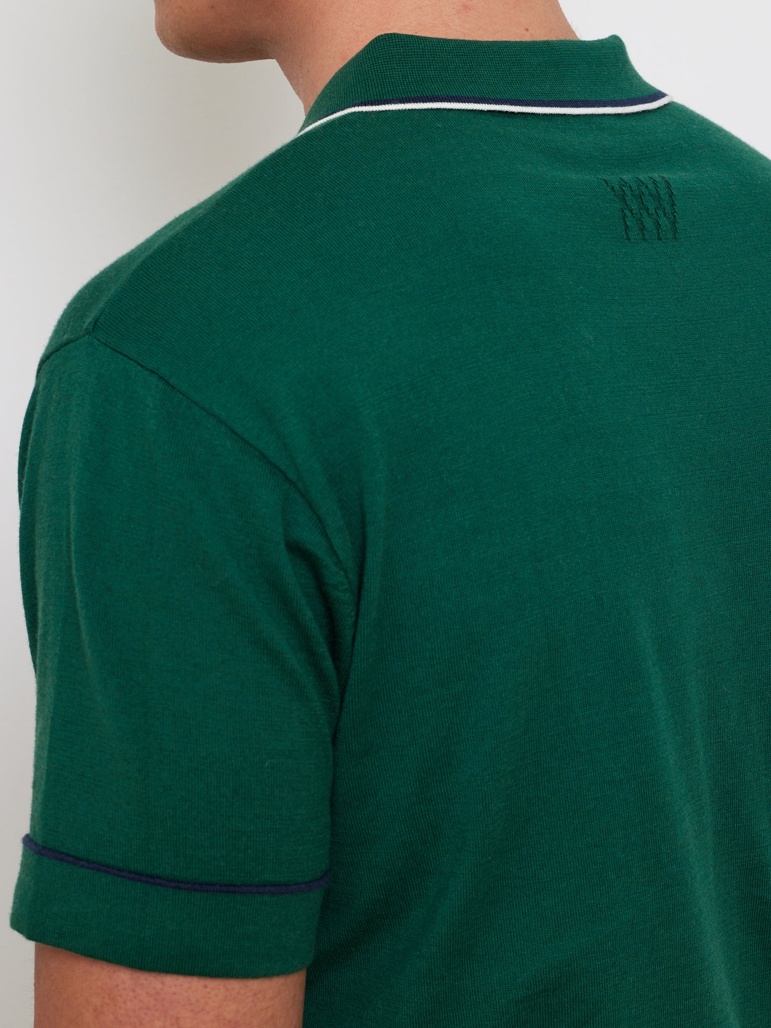 The person wears a We Norwegians Team Polo Men, crafted from breathable fabric in green with white and navy accents on the collar and sleeves. A subtle emblem adorns the back near the shoulder as they turn slightly away from the camera.