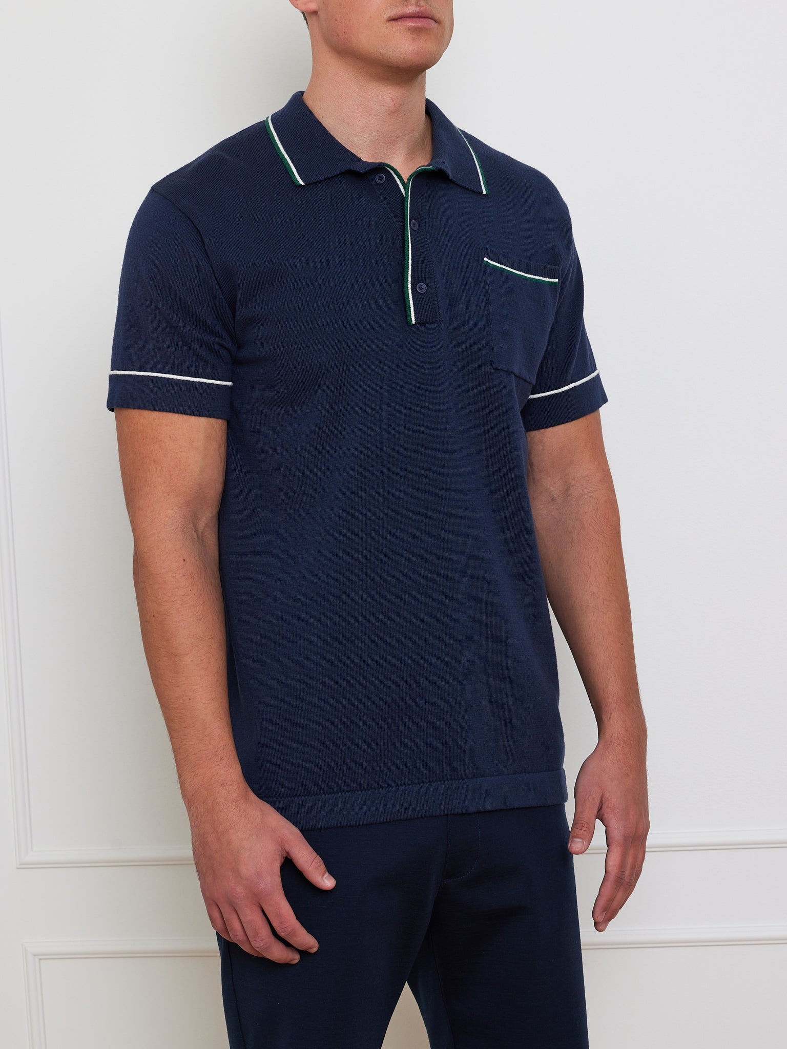 A person stands against a light wall wearing the We Norwegians Team Polo Men made from a breathable merino-cotton blend. This navy blue designer polo features contrasting white and green trim on the collar and sleeves, stylishly paired with navy pants for added comfort.