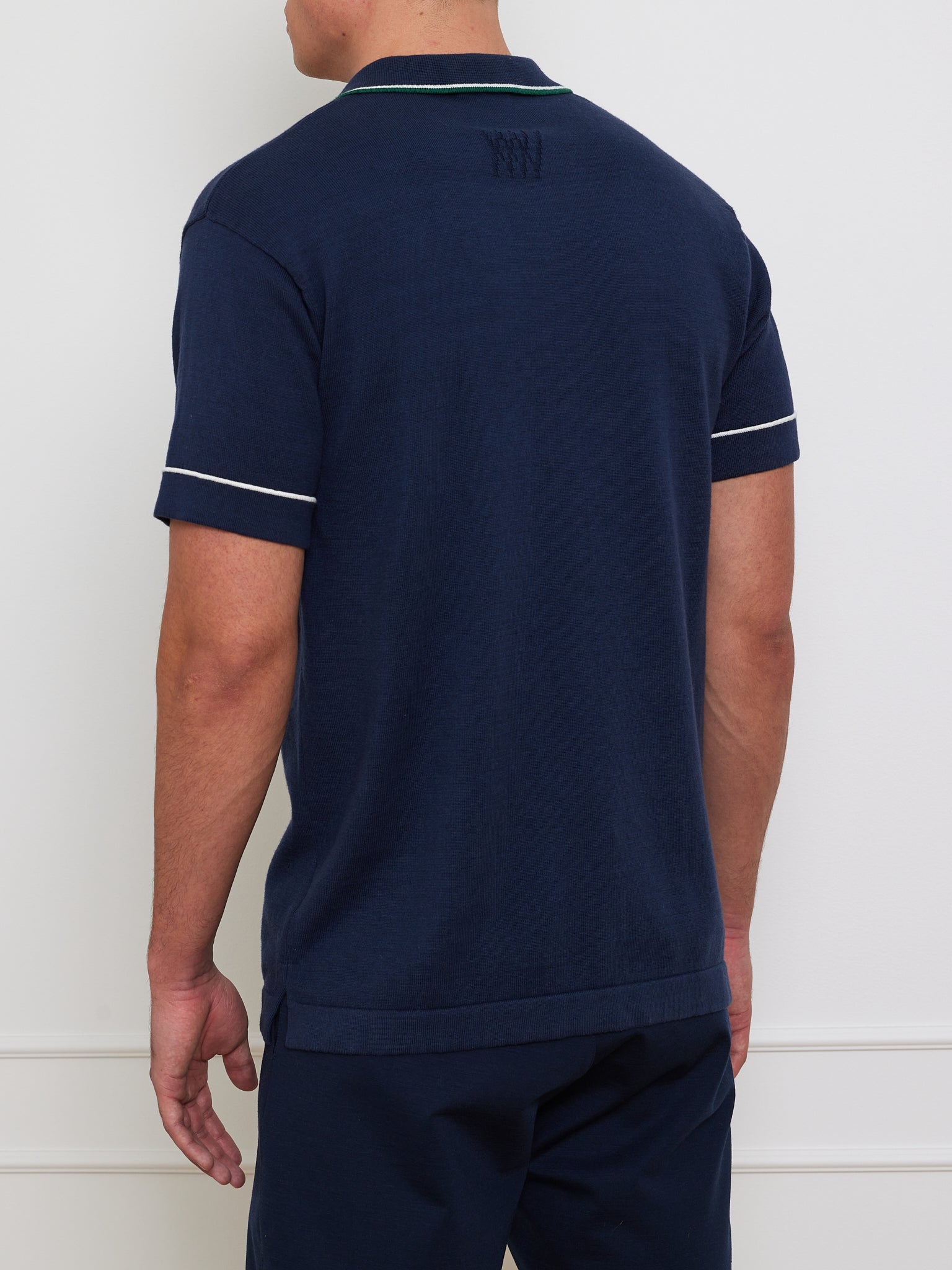 A person faces away from the camera, dressed in a dark blue Team Polo Men by We Norwegians, featuring white sleeve and collar trim. The untucked bottom of this merino-cotton blend stands out against the plain white wall.