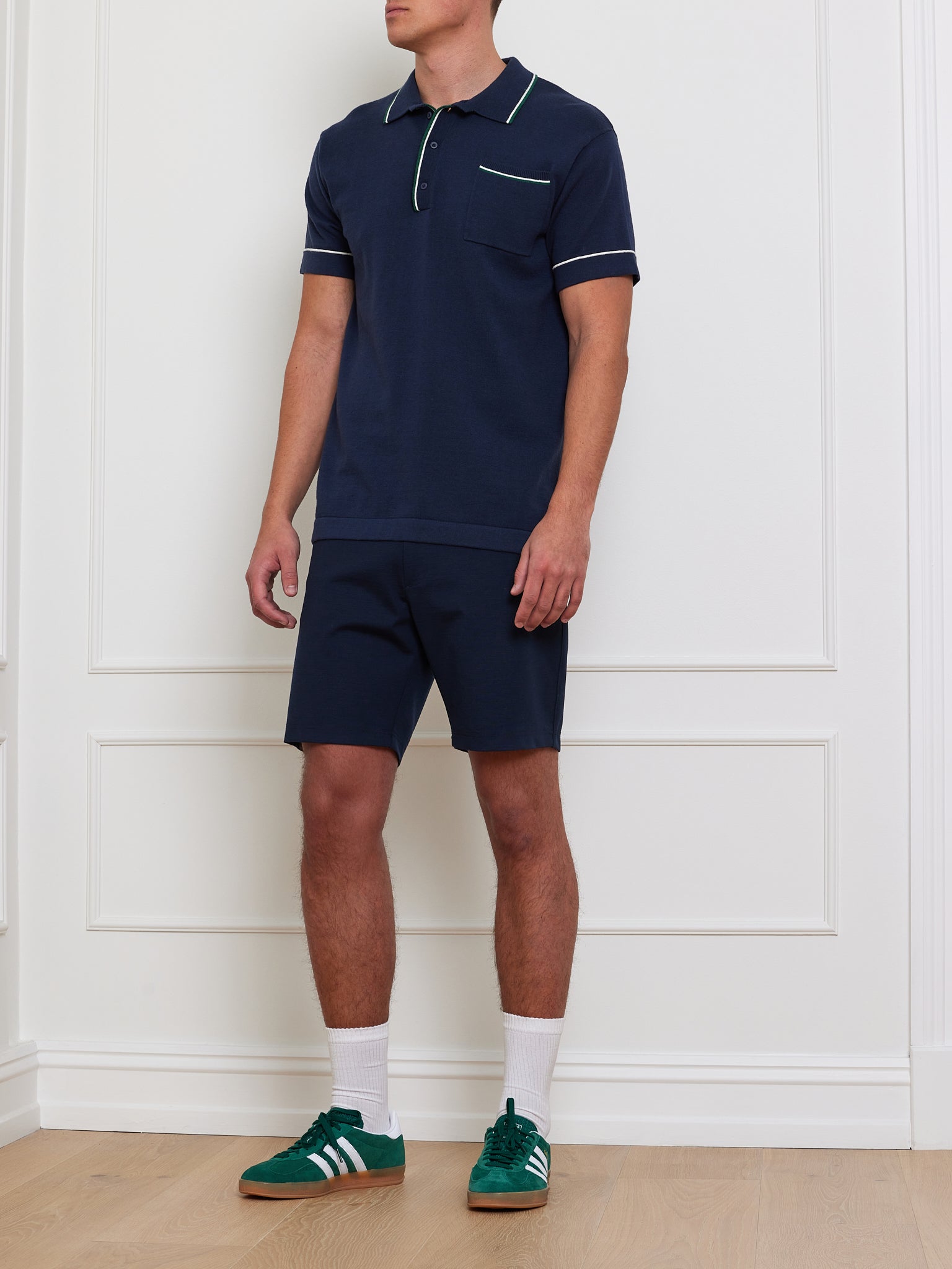 Wearing a We Norwegians Team Polo Men navy shirt and matching shorts, along with white socks and green sneakers with white stripes, a person stands against a white wall. Their face is not visible.