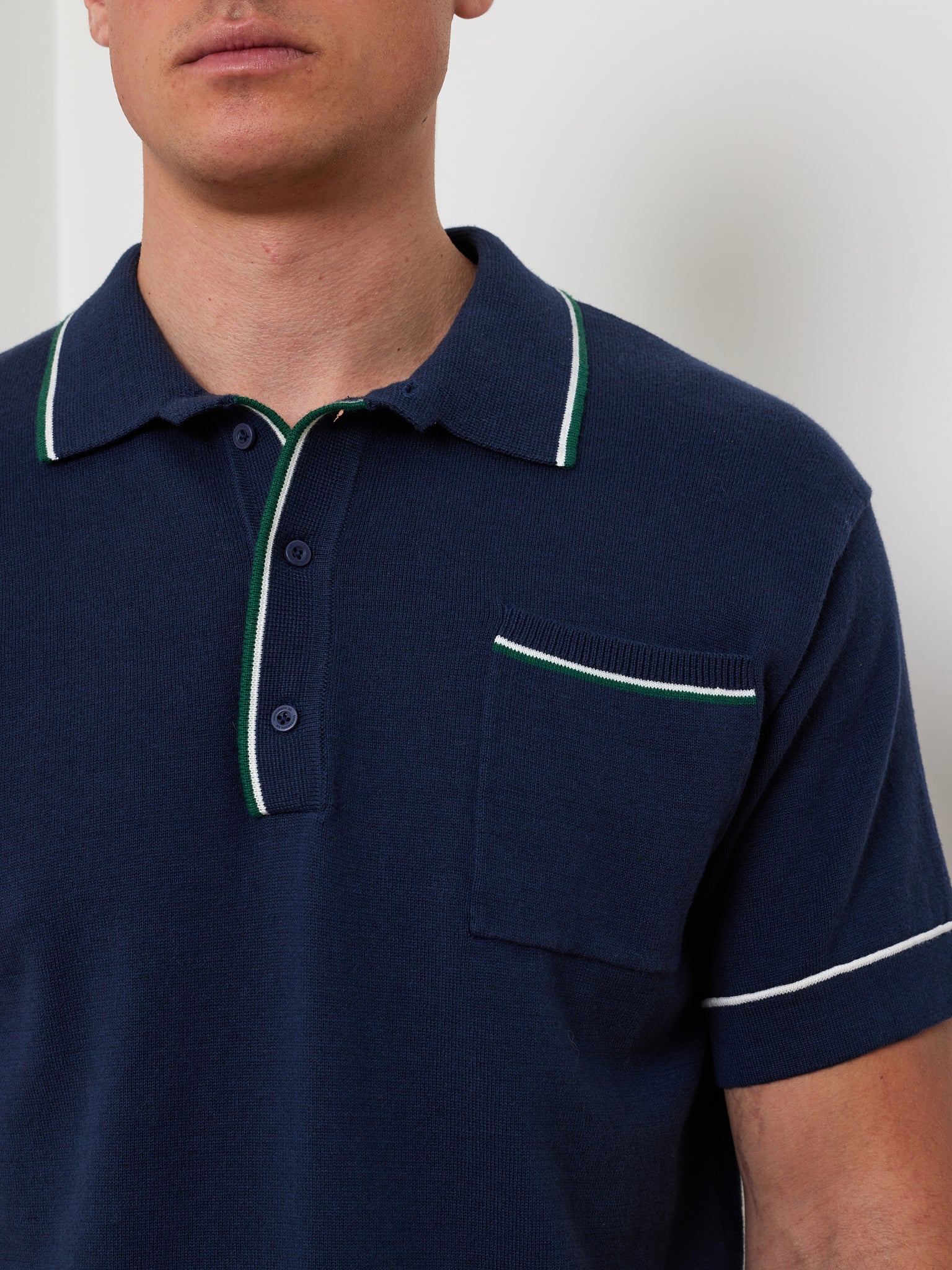 A person wears the "Team Polo Men" by We Norwegians, featuring a merino-cotton blend fabric in navy blue with white and green trim on the collar, placket, and pocket. The shirt includes a front pocket with contrast piping, partially obscuring their face.