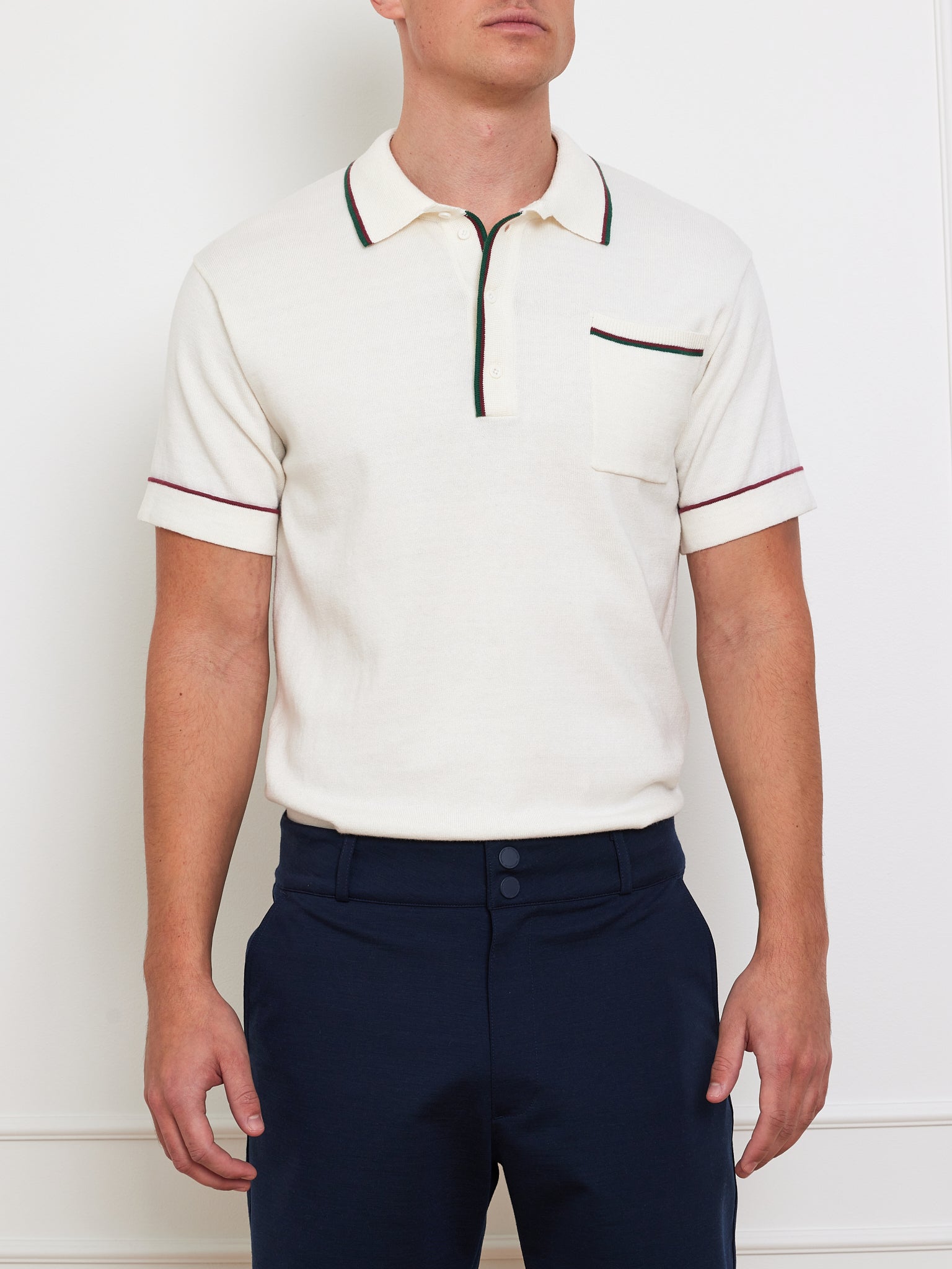 A person wears a white Team Polo Men by We Norwegians, featuring short sleeves, a ribbed collar, and black and red contrast piping with a front pocket. They're paired with dark blue pants against a plain white wall.