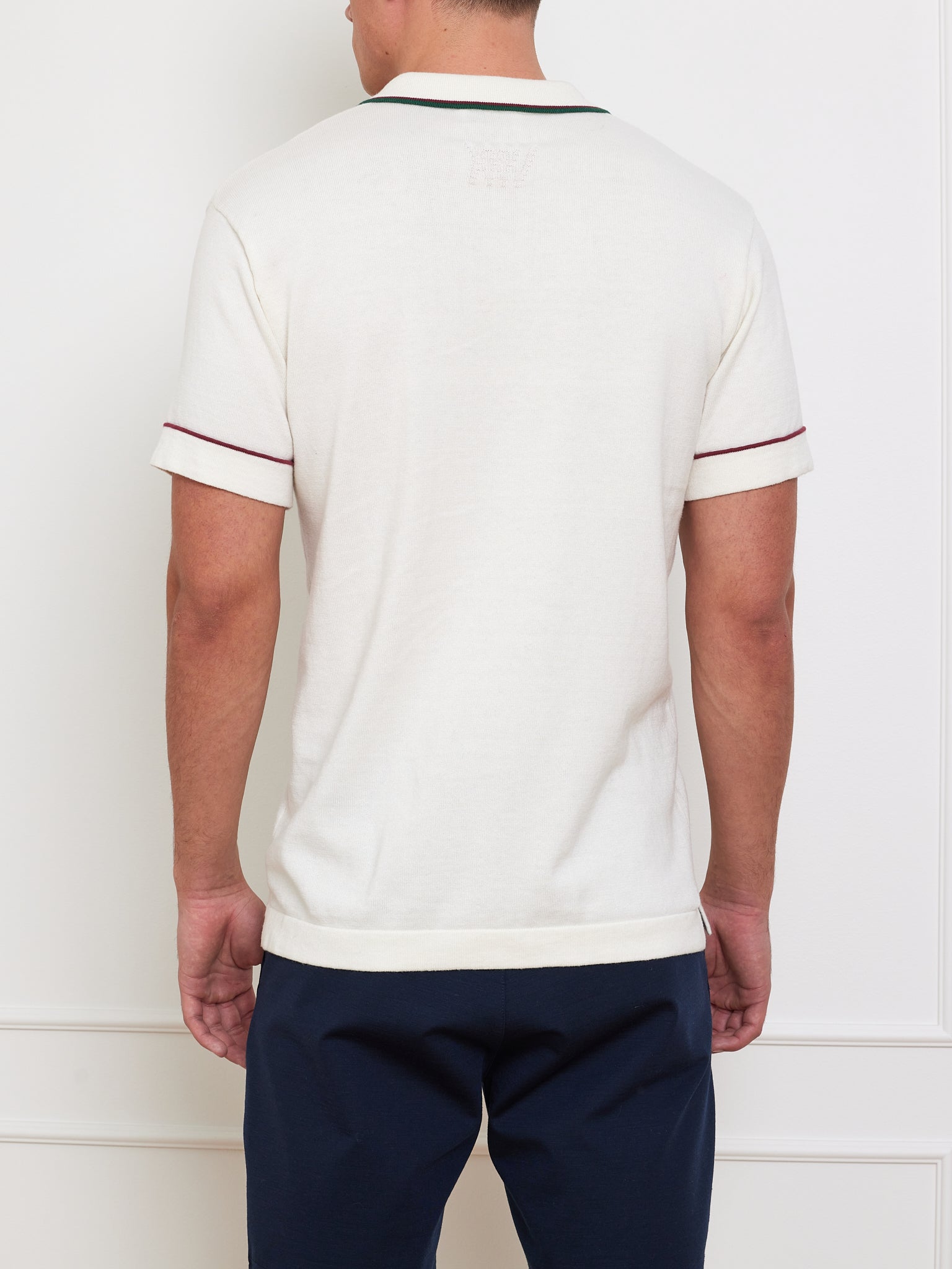 A person stands with their back to the camera in a We Norwegians Team Polo Men, featuring short sleeves, cream color, contrast piping on the sleeves, and a ribbed collar. They wear dark pants against a plain white wall with subtle paneling.