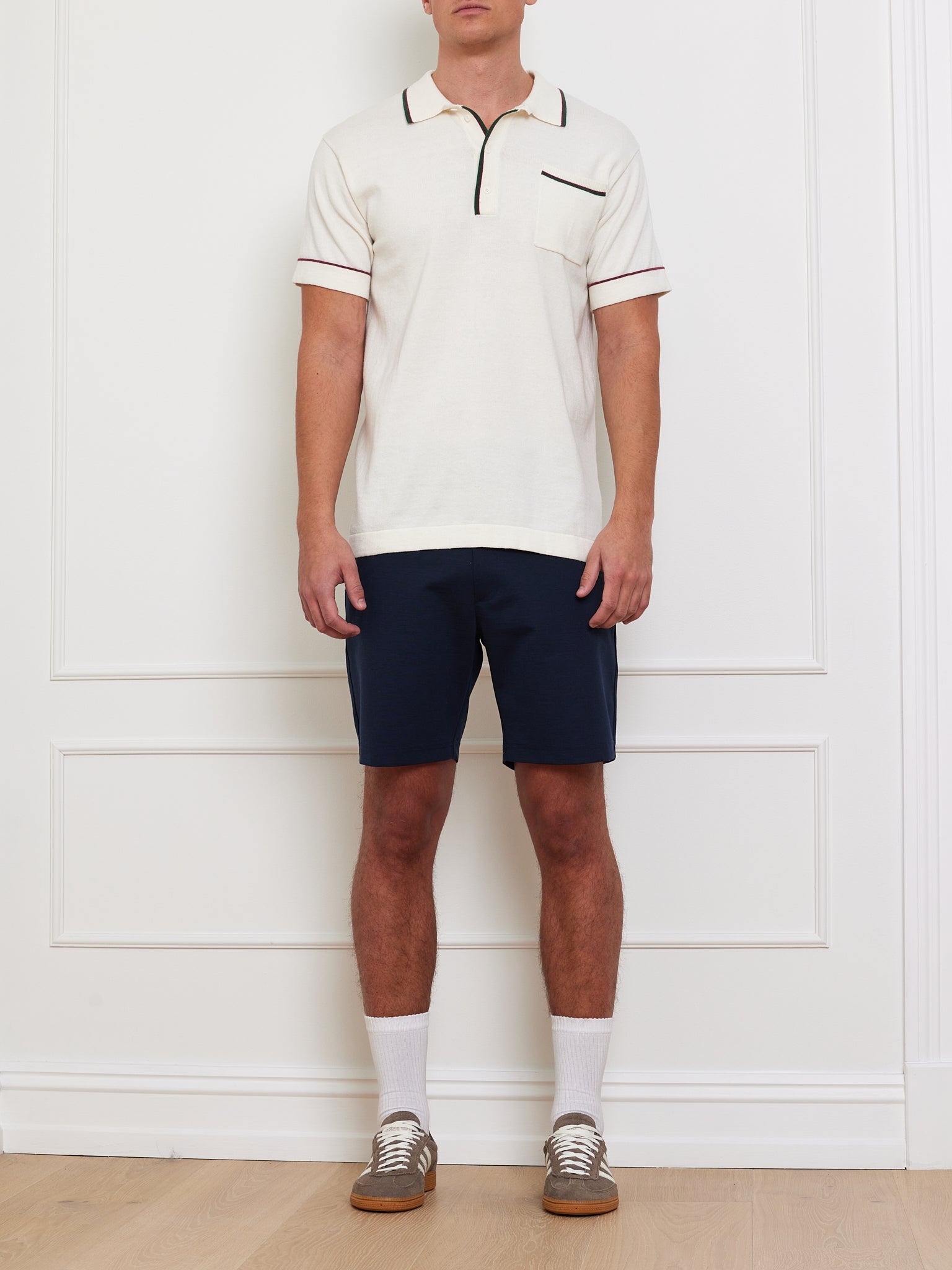 A person wearing a We Norwegians Team Polo Men shirt with black contrast piping, navy blue shorts, white socks, and brown sneakers stands indoors against a white wall with decorative trim on a light wood floor.