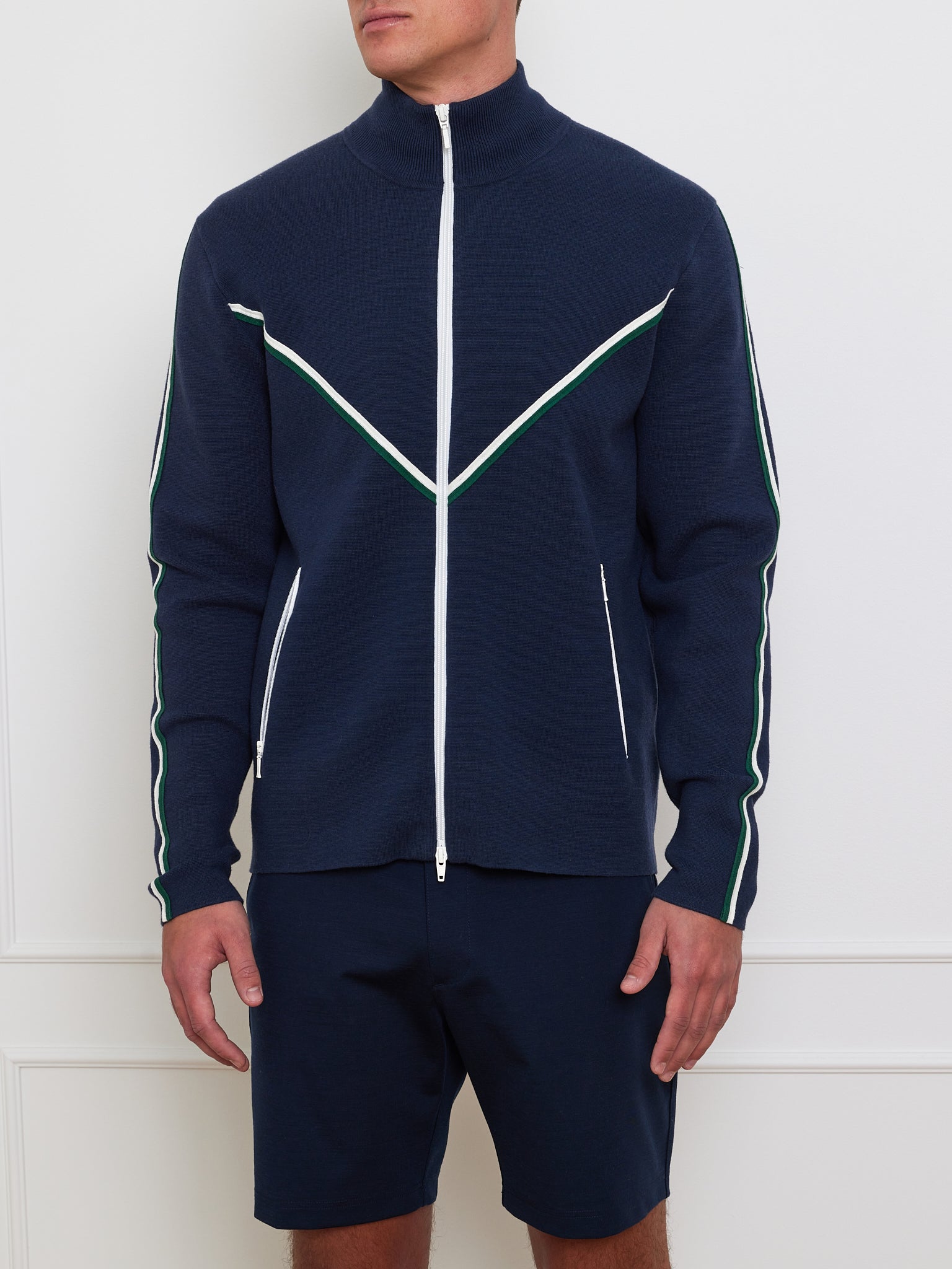 A person models the "Team Jacket Men" by We Norwegians, showcasing its retro design with a navy cotton-wool blend, white and green sleeve stripes, and chevron chest piping. They pair it with navy shorts while standing against a white wall.