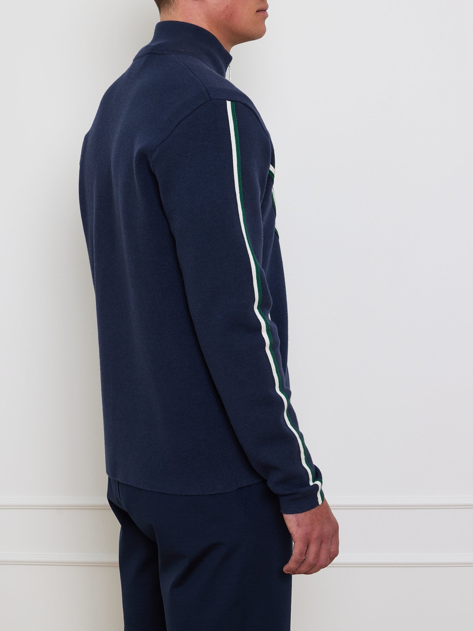 A person is sideways against a light background, wearing We Norwegians' Team Jacket Men, a navy retro track jacket with white and green sleeve stripes and chevron piping. Their hands rest at their sides.