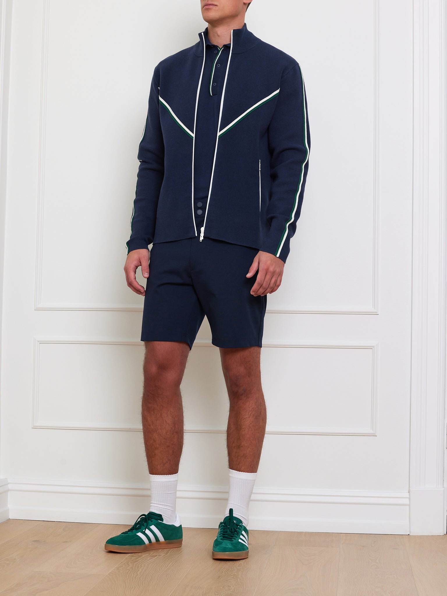 A man sports the We Norwegians Team Jacket Men, adorned with white accents, over a matching polo and shorts. He pairs it with white socks and green sneakers featuring white stripes. The ensemble with chevron piping stands against a minimalist setting of a white wall and wooden floor.