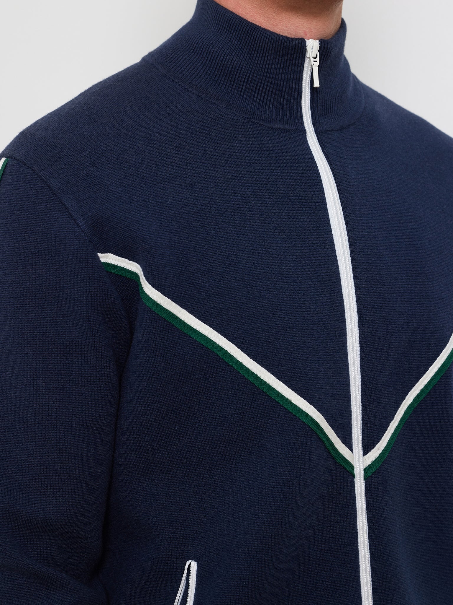 Sporting the Team Jacket Men by We Norwegians, a person wears a navy zip-up with chevron piping on the chest, crafted from a cozy cotton-wool blend. White and green stripes form a V pattern on the midsection, while only part of their face is visible against a plain backdrop.