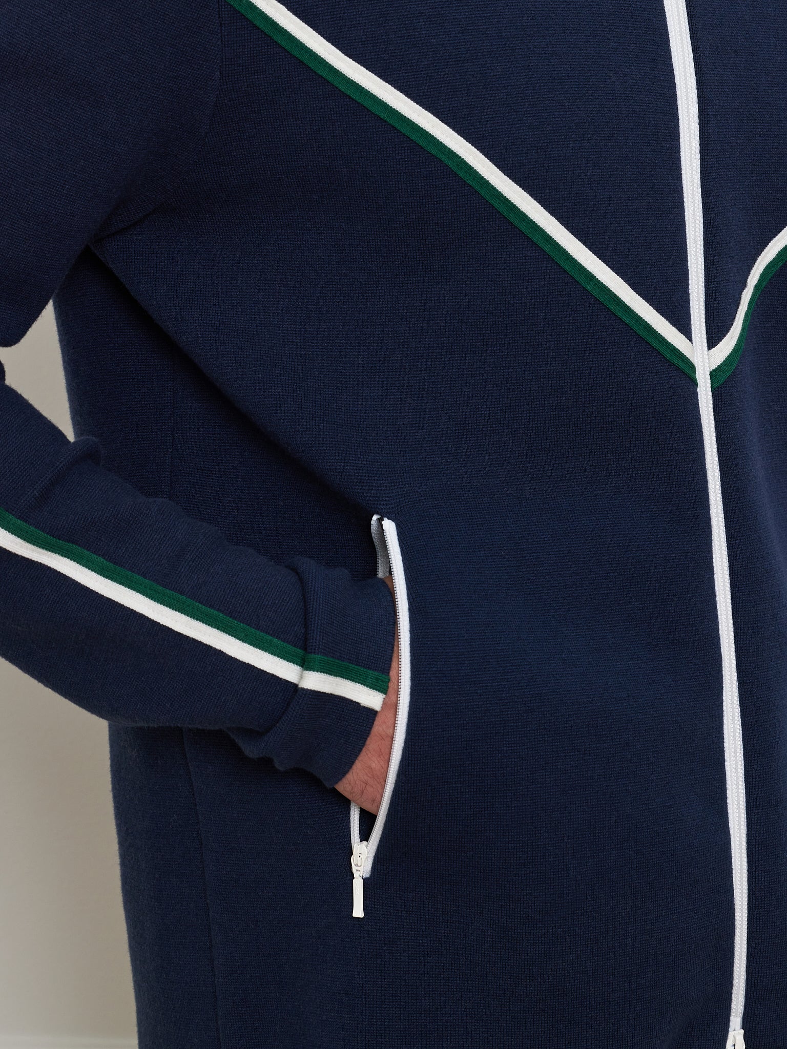 A person wears the We Norwegians Team Jacket Men in navy blue, featuring white and green diagonal stripes, pockets, and stylish chevron piping. One hand rests casually in a pocket.