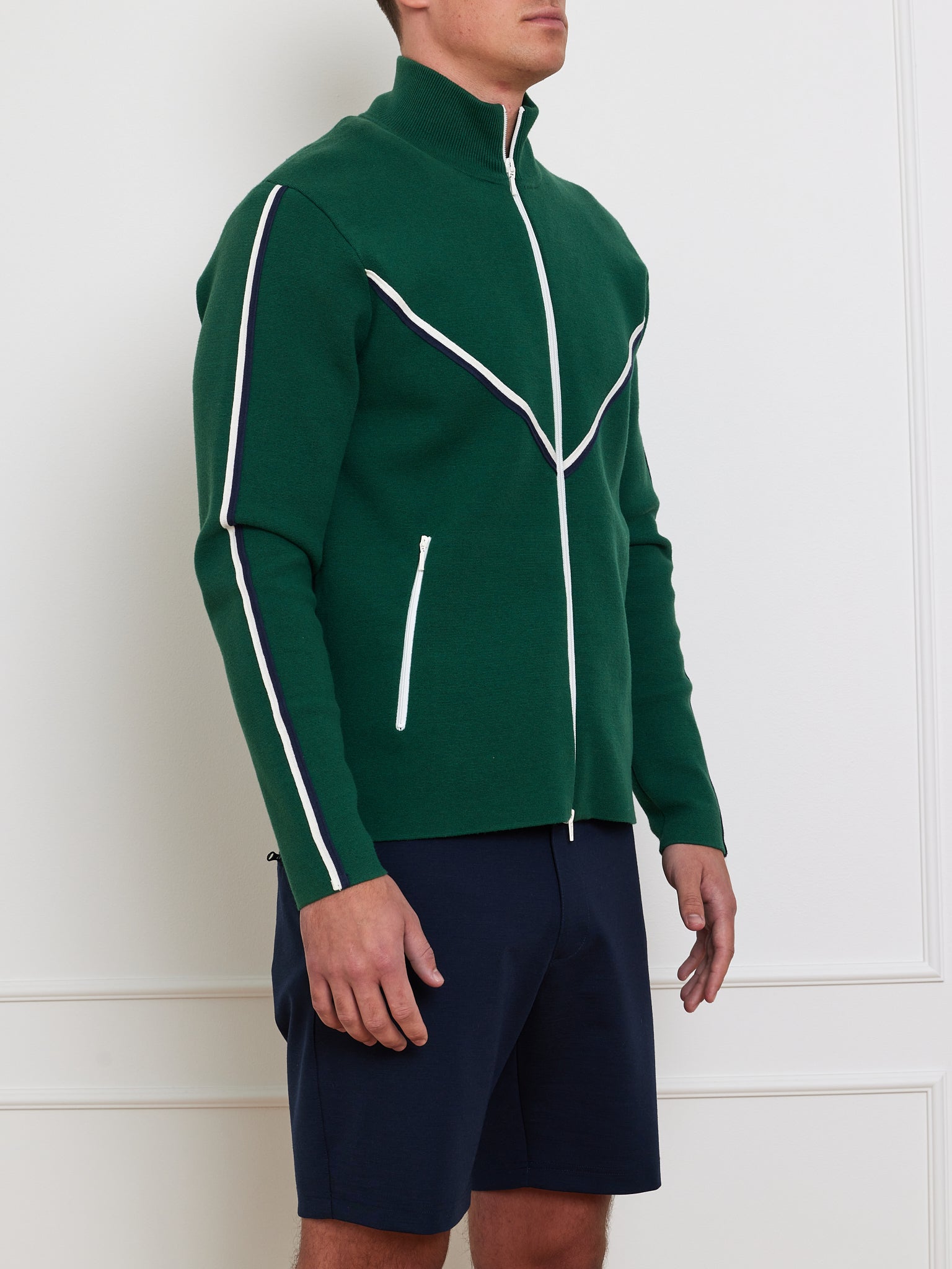 A man wears the We Norwegians Team Jacket Men, a retro track jacket in vibrant green with white and black trim on the sleeves and shoulders. It features vertical zippered pockets for a nostalgic vibe. Paired with navy blue shorts, he stands against a crisp white wall.