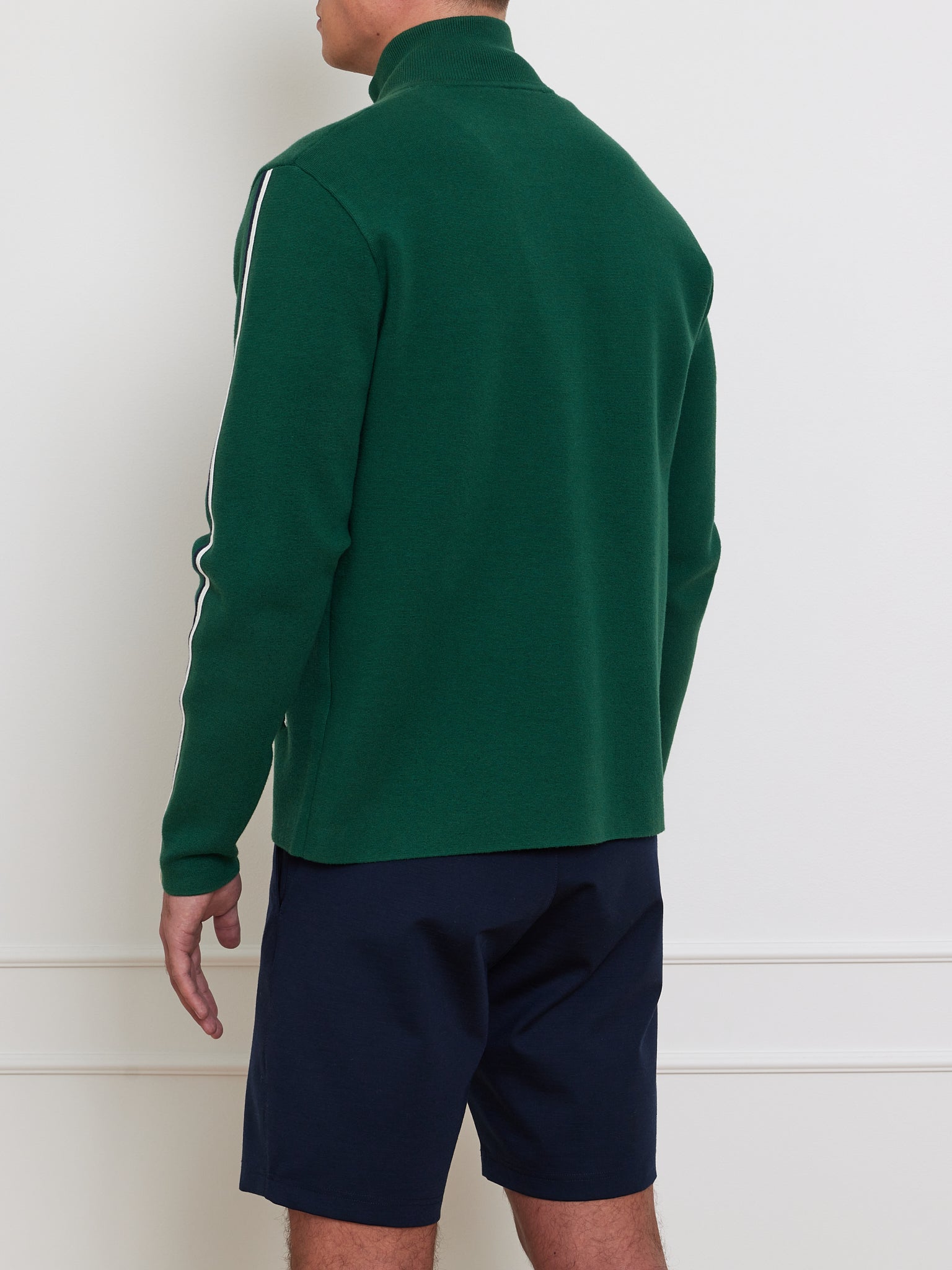 A person wearing a We Norwegians Team Jacket Men, featuring retro chevron piping and navy shorts, stands with their back to the camera. The jacket's green fabric boasts white sleeve stripes, set against a plain white wall with molding.
