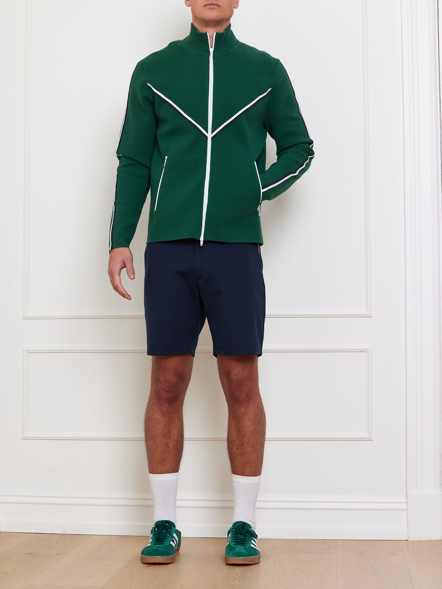 A person stands against a white wall wearing the We Norwegians Team Jacket Men, featuring white chevron piping, and navy shorts. They also have long white socks and green sneakers. The light-colored wood floor complements the sporty look.