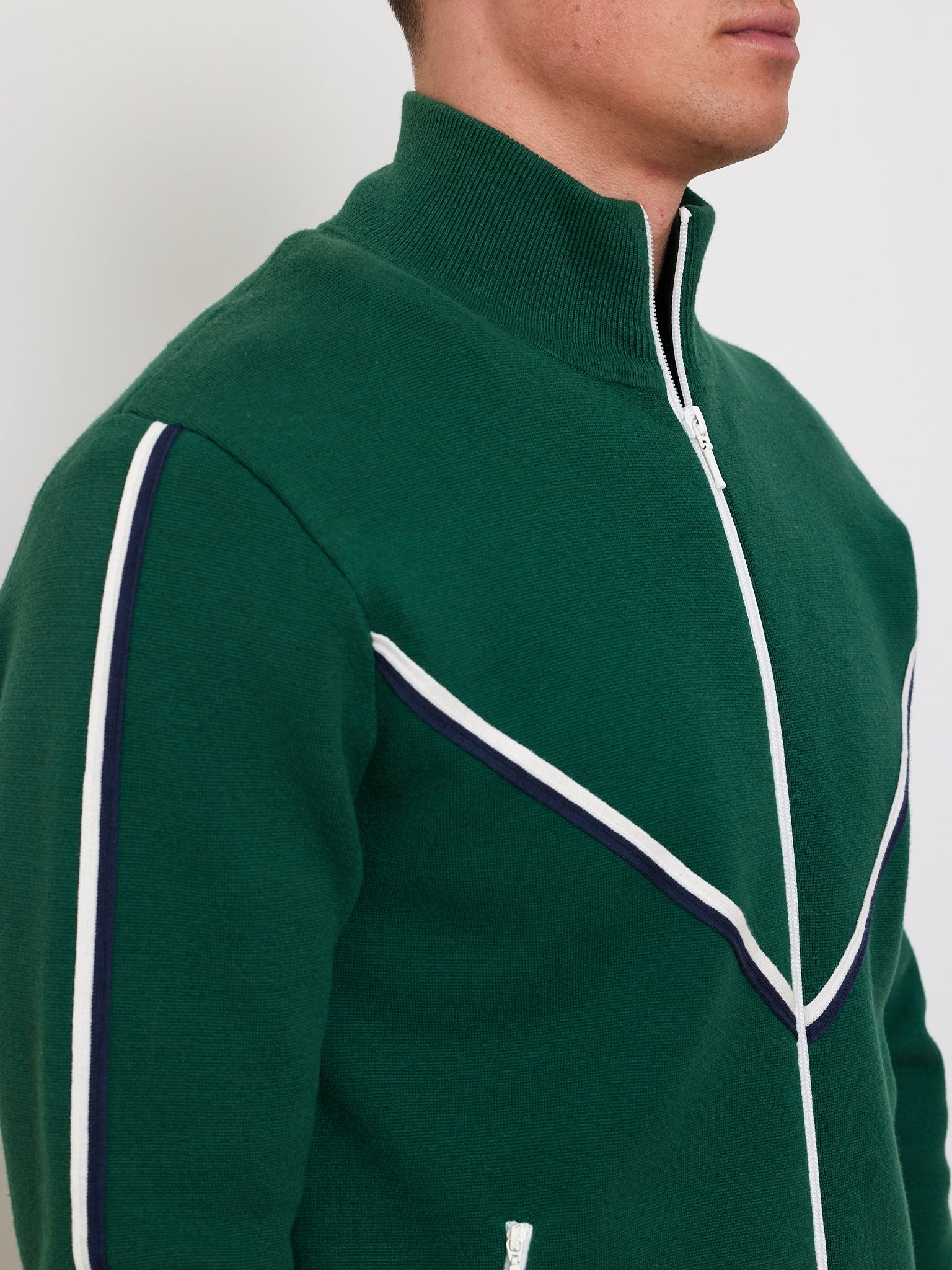 A man models the Team Jacket Men by We Norwegians in green, styled with a retro track vibe featuring white and navy chevron piping. Made from a cotton-wool blend, it has a high collar and white zipper details, captured from the side against a plain backdrop.