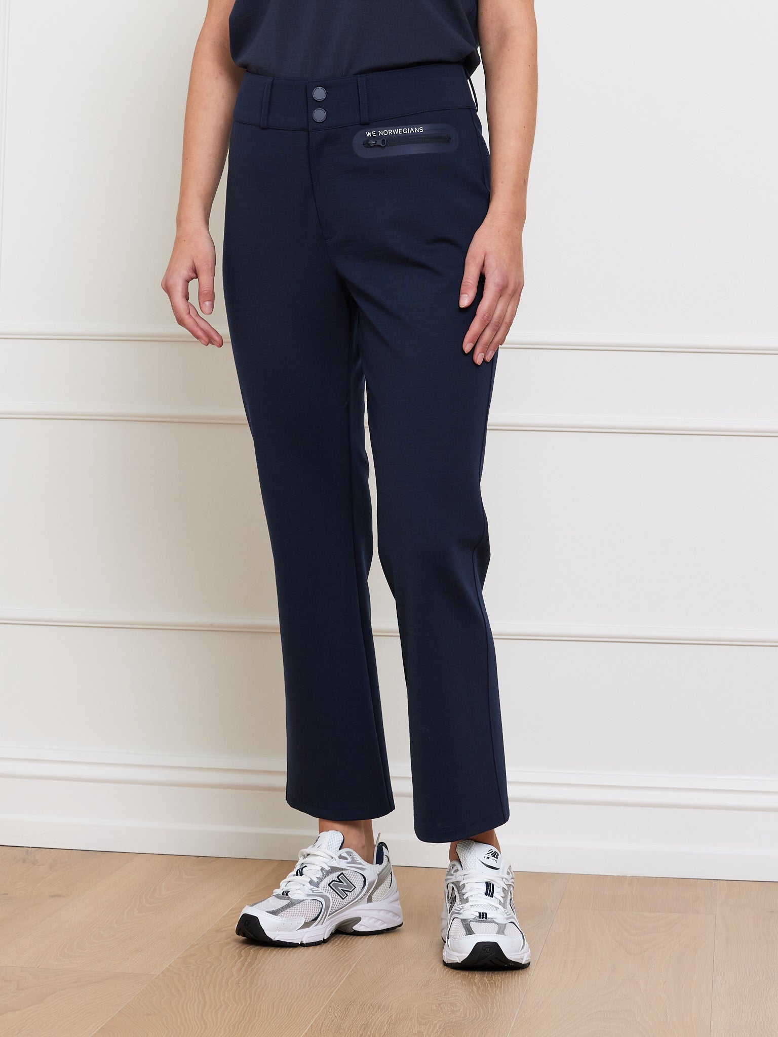 A person stands against a white wall in navy blue Ace Pant Women by We Norwegians, paired with a dark top and white sneakers. The trousers include waist buttons, subtle pockets, and offer 4-way stretch for comfort. The floor is light-colored wood.