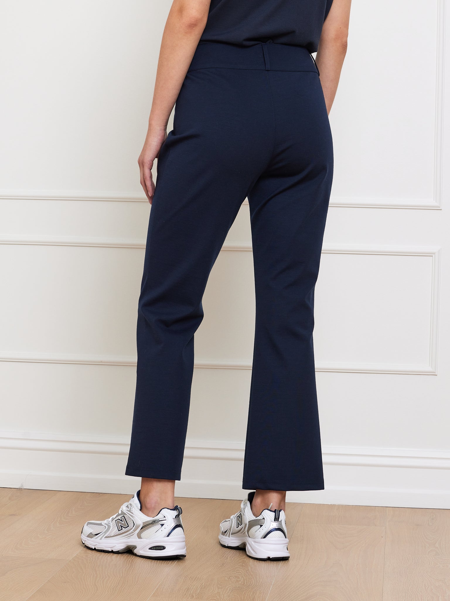 Rear view of a person in We Norwegians' Ace Pant Women in dark blue with Tencel™ fabric, paired with white sneakers on a wooden floor. A black shirt complements the relaxed style against a white paneled wall backdrop.