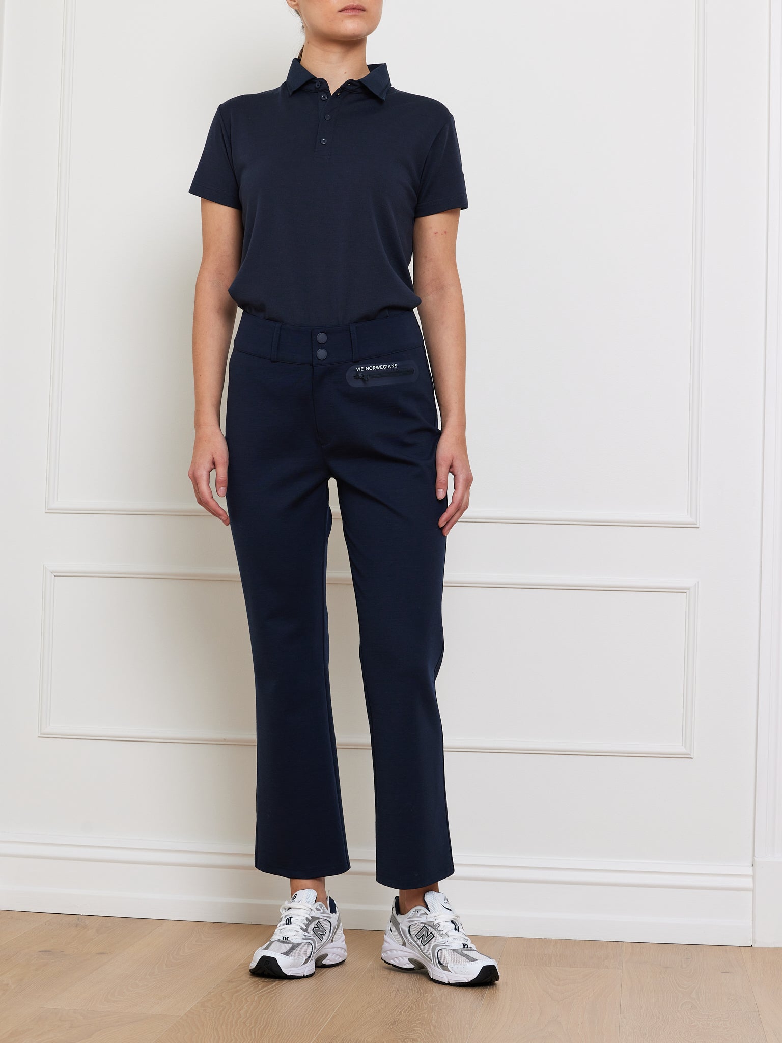 A person in a black polo shirt, navy women's Ace Pant by We Norwegians with Tencel™, and white athletic shoes stands against a white wall with decorative molding on a light wooden floor.
