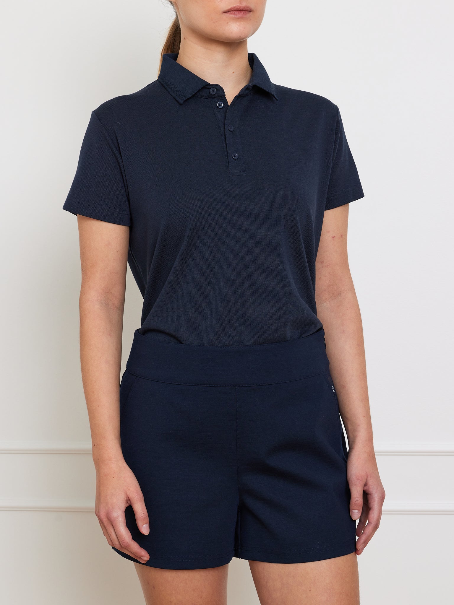 A person is wearing a dark navy polo shirt and We Norwegians' Ace Shorts Women, combining comfort with style in a classic collared design and short sleeves, set against a plain white wall backdrop.