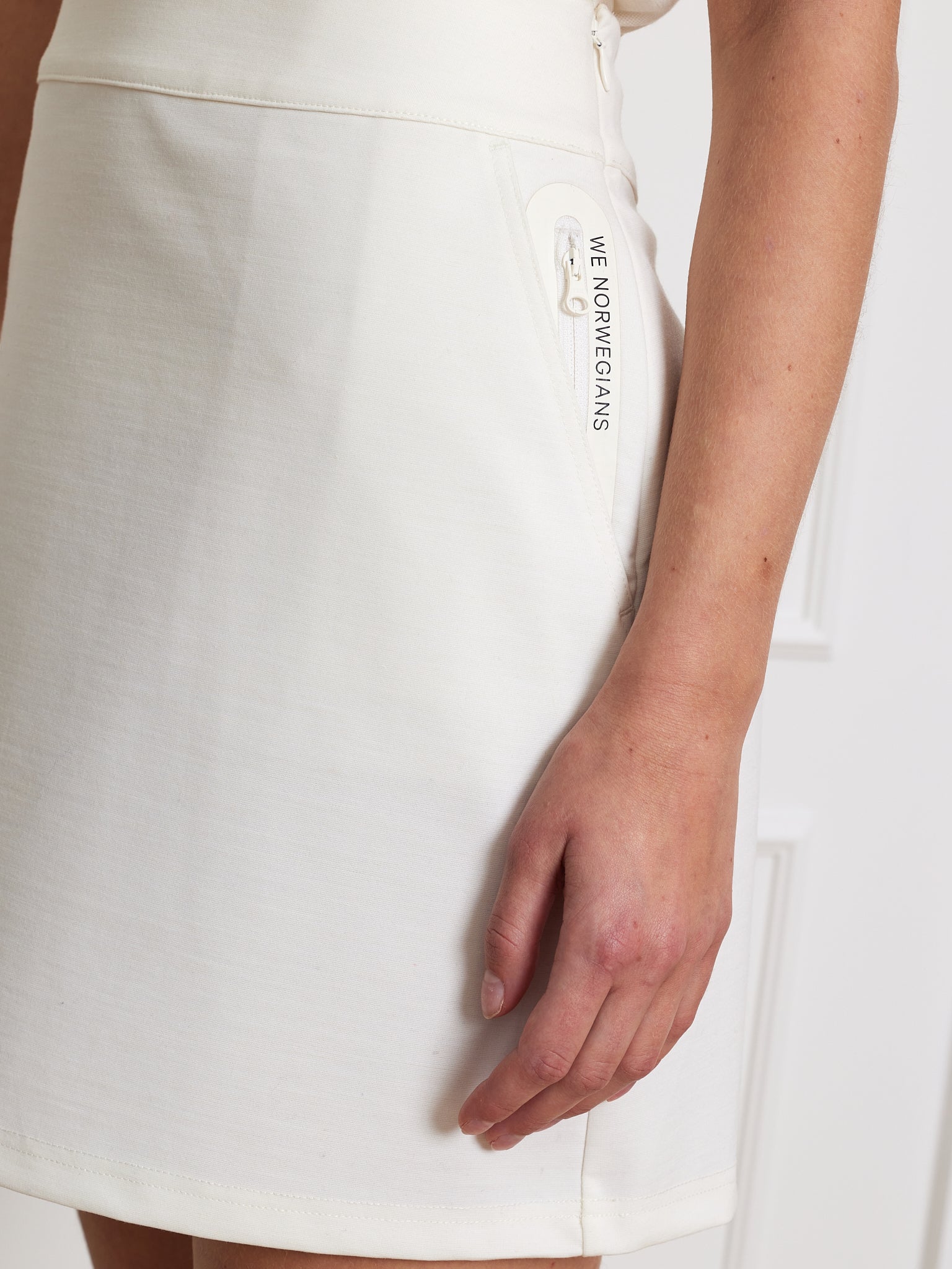 Close-up of a person wearing the Ace Skirt Women, a high-waist wool blend with a side zipper. The brand "We Norwegians" is printed near the zipper. Made from sustainable fabric, it is set against a white background with the person's hand by their side.