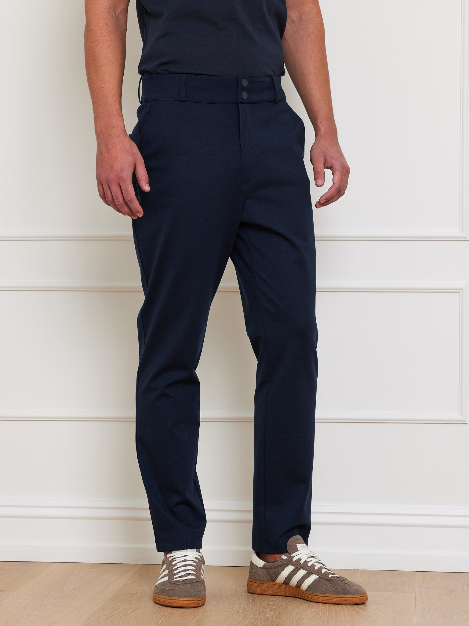 A man indoors stands against a white-paneled wall, wearing We Norwegians' Ace Pant Men with Tencel™ Lyocell in navy and a black t-shirt, paired with brown and white sneakers. His face remains unseen.