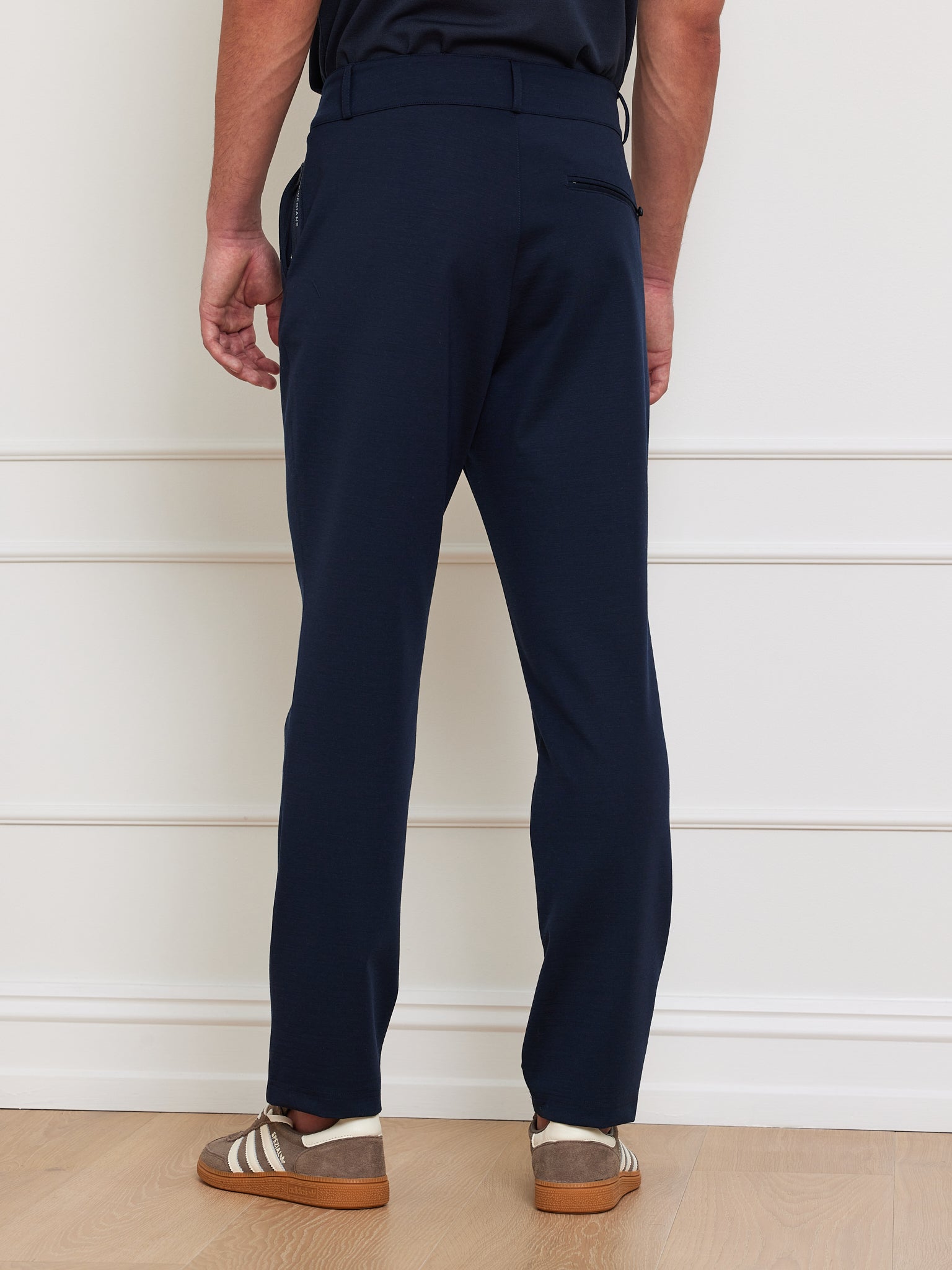 A person wearing We Norwegians' Ace Pant Men in navy blue, crafted from Tencel™ Lyocell with a tapered fit and 4-way stretch, is complemented by brown sneakers with white stripes. The pants include a back pocket and are showcased on a wooden floor with a white wall backdrop.