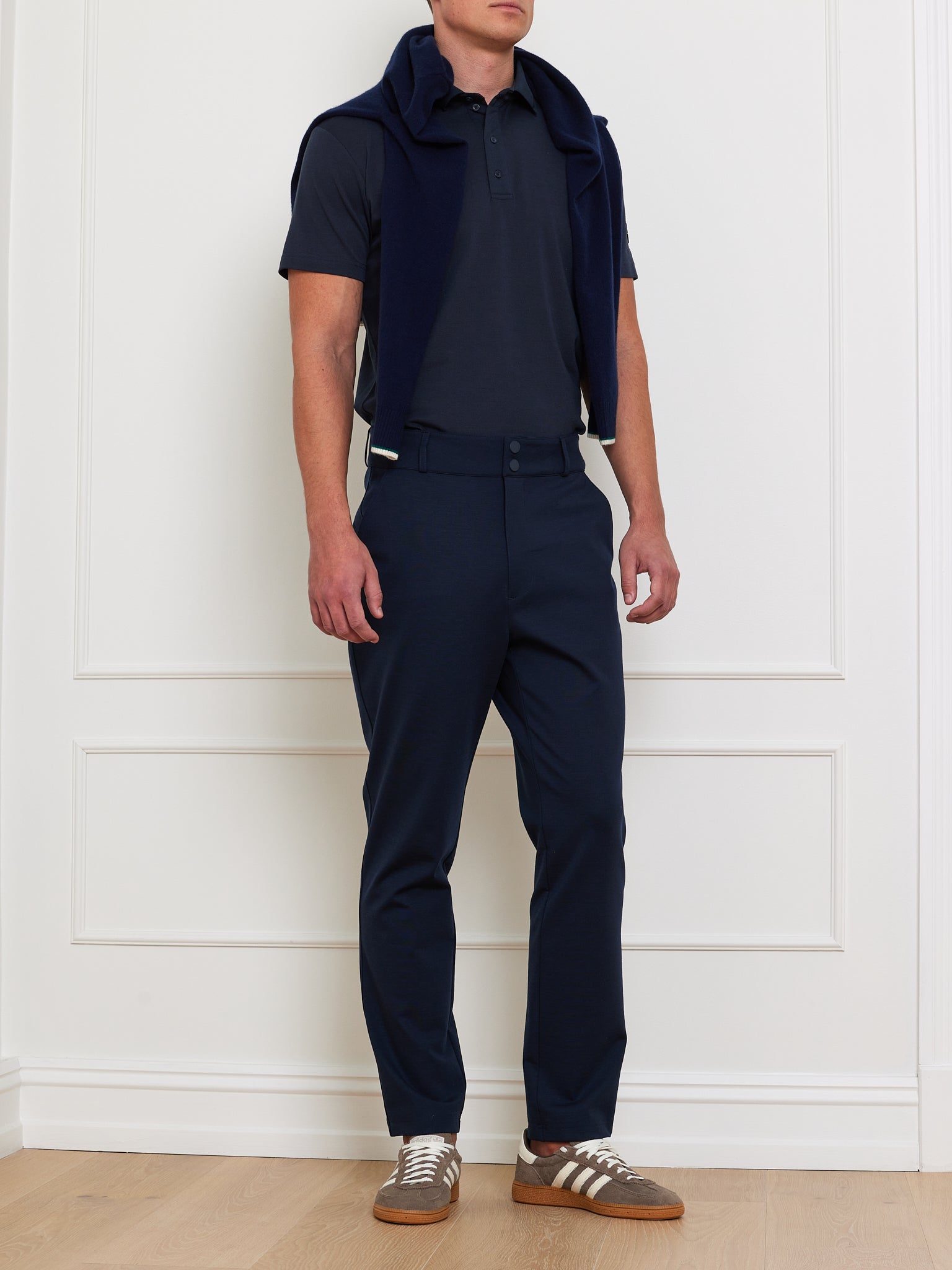 A person in a We Norwegians navy polo shirt and Ace Pant Men stands against a white wall, with a navy sweater draped over their shoulders. They wear brown and gray sneakers, all set in a minimal room with wooden flooring, showcasing the outfit's 4-way stretch fabric made from Tencel™ Lyocell.