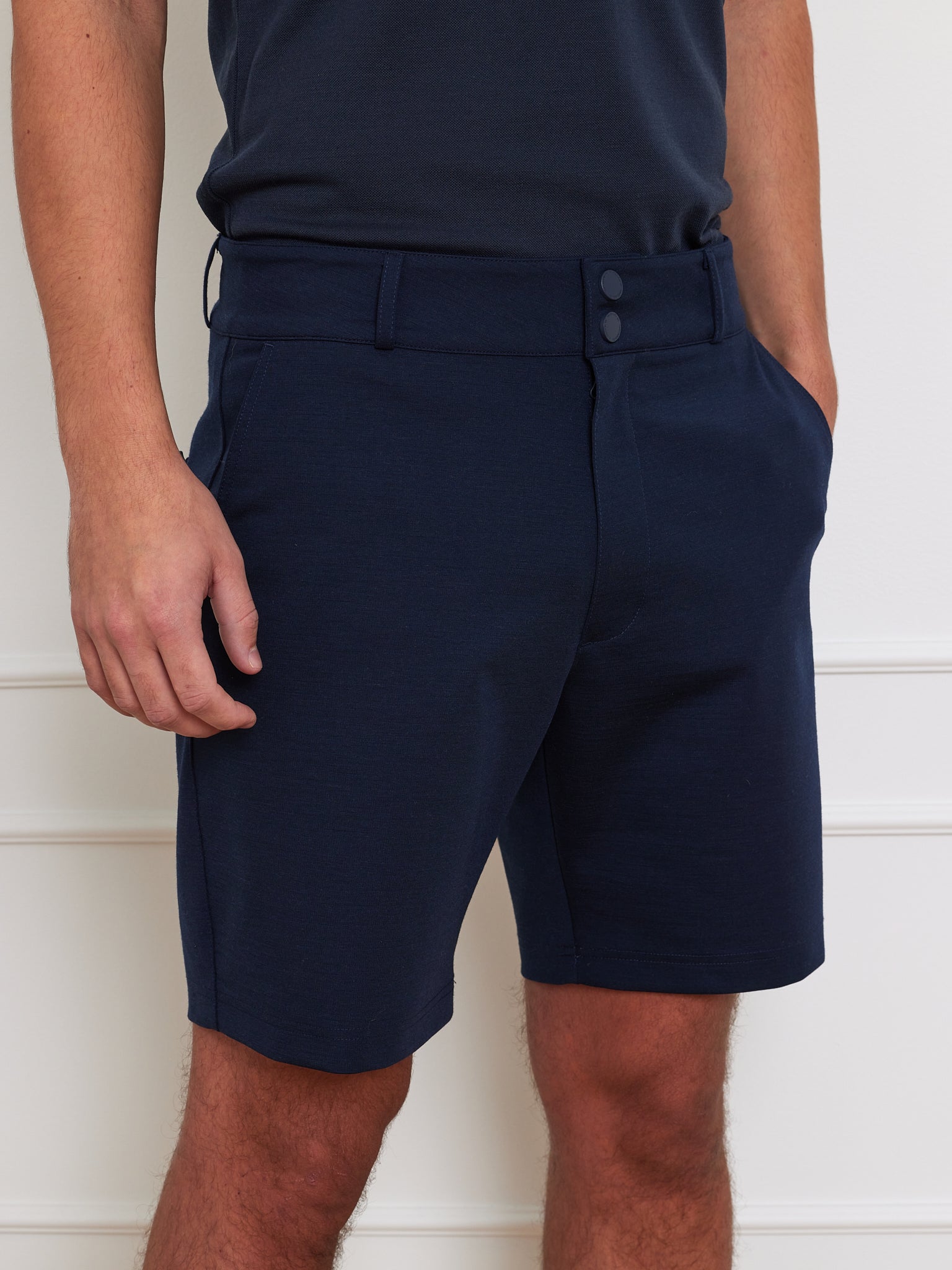 A person wearing We Norwegians Ace Shorts Men in navy blue, crafted from Tencel™ with button details and 4-way stretch fabric, stands against a white wall. The shorts feature pockets where the person's hands are casually placed, paired with a matching navy top.