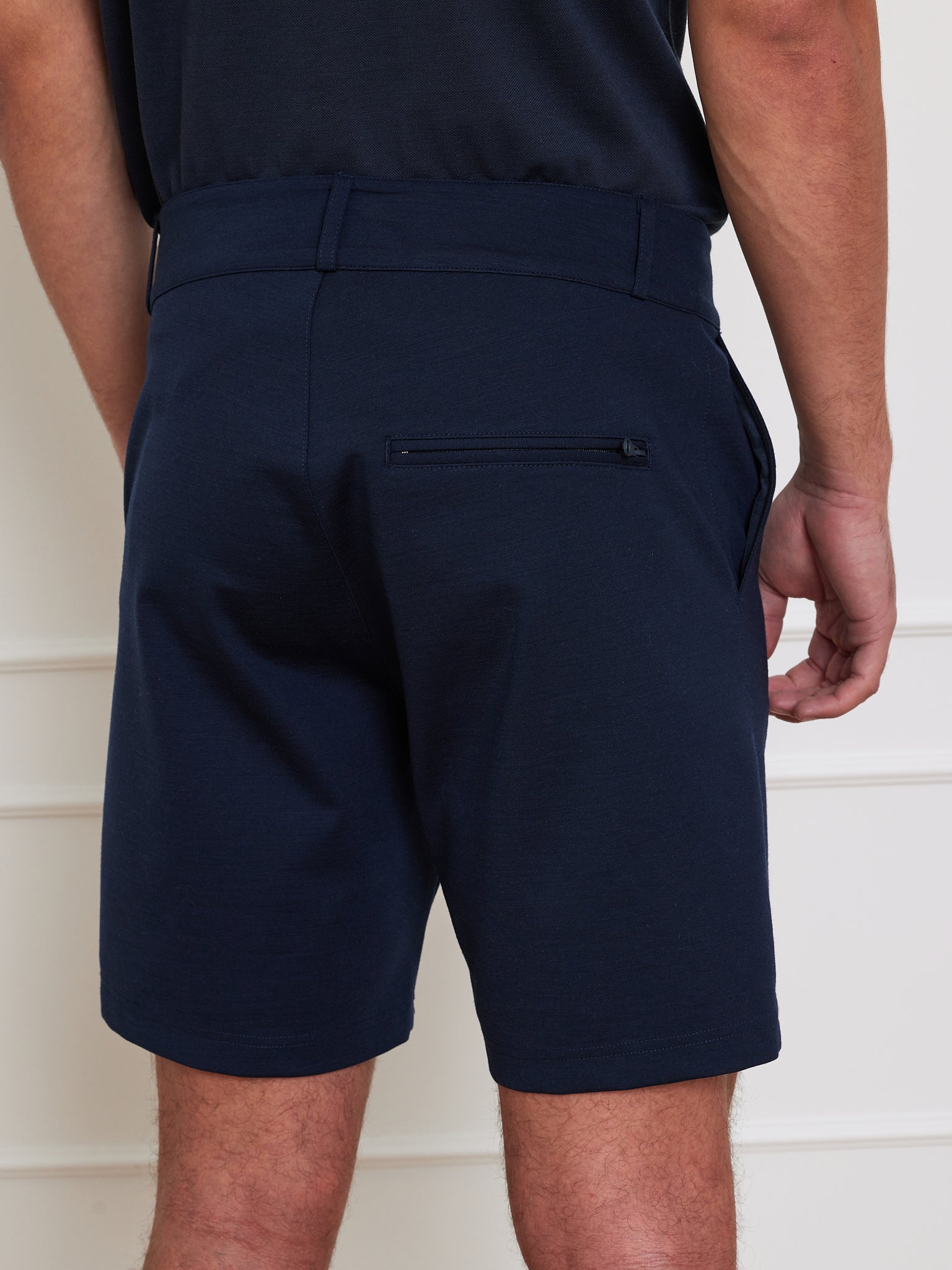 A person in We Norwegians' Ace Shorts Men, featuring a navy blue color and 4-way stretch with a zipped right pocket, stands against a light wall. Part of a black shirt is visible at the waist as their hands rest by their sides.