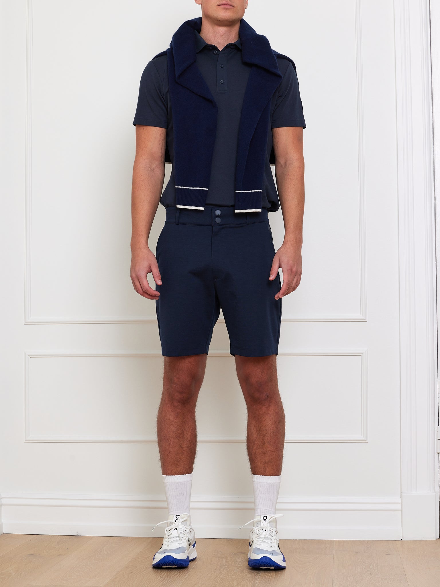 In a minimalistic room with light wood flooring, a person poses against a white paneled wall wearing We Norwegians' Ace Shorts Men and navy polo, paired with white sneakers featuring blue accents and a premium wool navy sweater draped over their shoulders.
