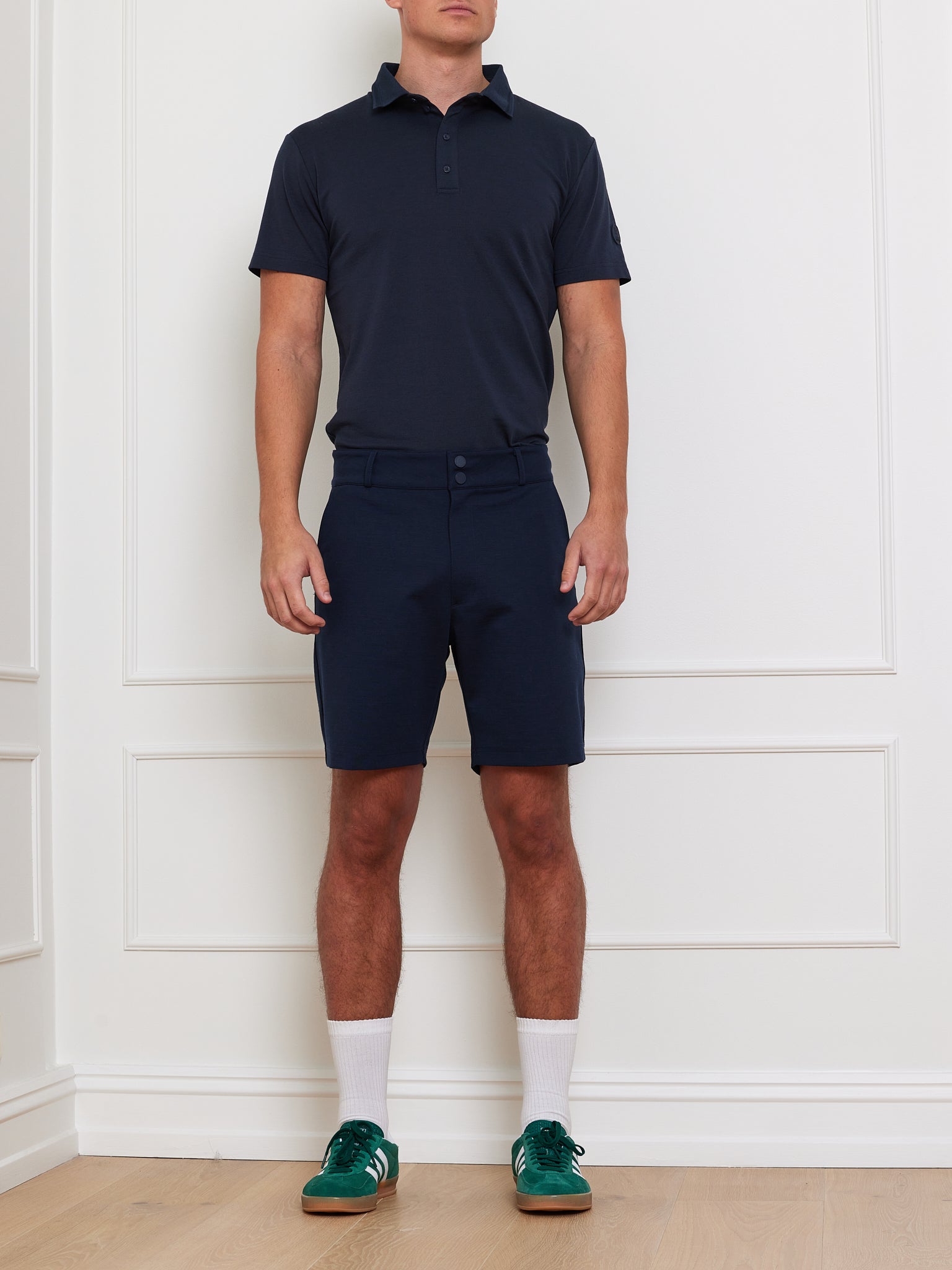 In front of a white wall, a person wears We Norwegians' Ace Shorts Men in dark blue, made with breathable Tencel™. The look includes matching polo, white socks, and dark green sneakers; their head is not visible.