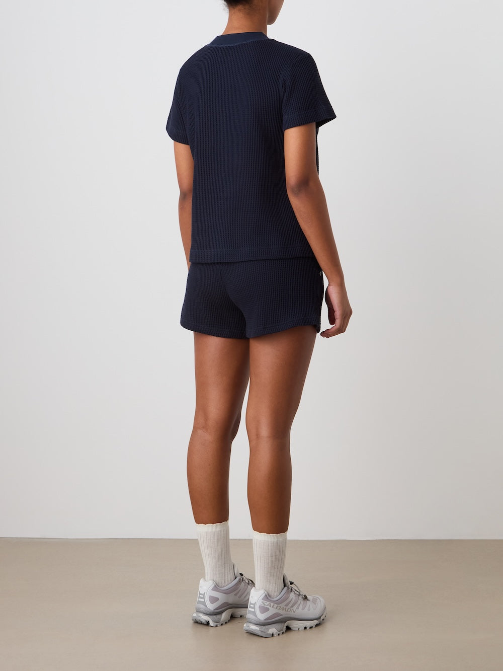 A person stands indoors facing away, wearing the We Norwegians Waffle Tee Women in navy with matching shorts, plus white crew socks and gray sneakers, against a plain light-colored wall and floor.