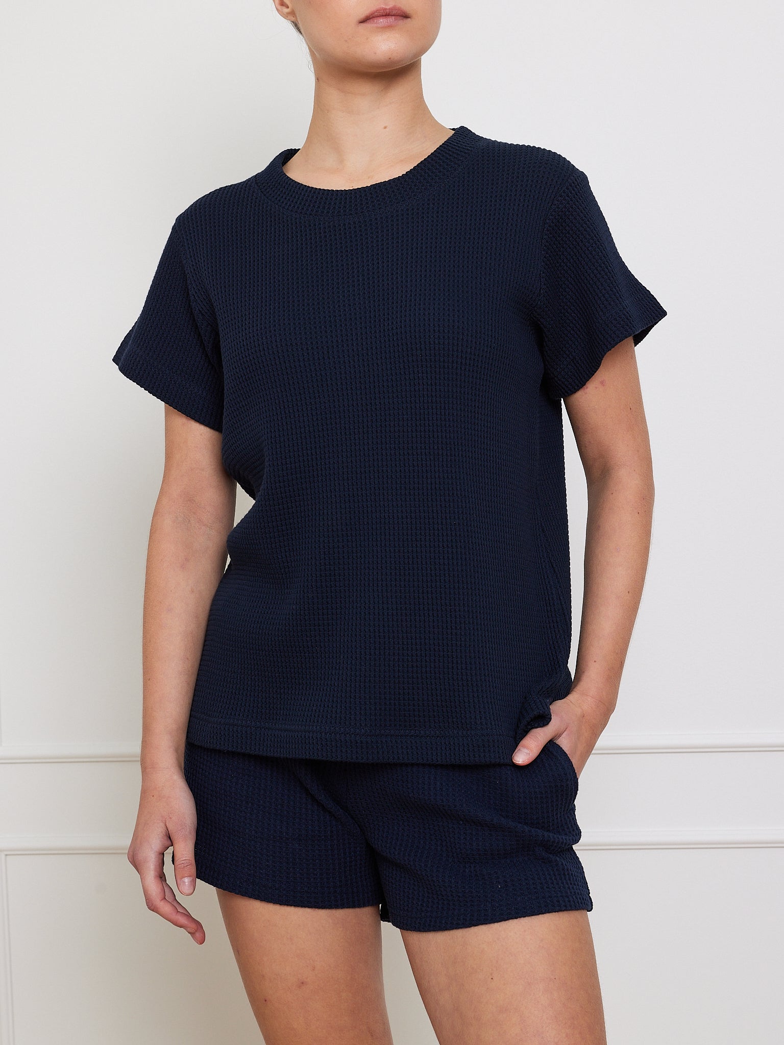 A person in a dark blue We Norwegians Waffle Tee Women and matching shorts stands against a plain light wall, with their left hand partially tucked into the shorts pocket, highlighting this classic wardrobe essential.