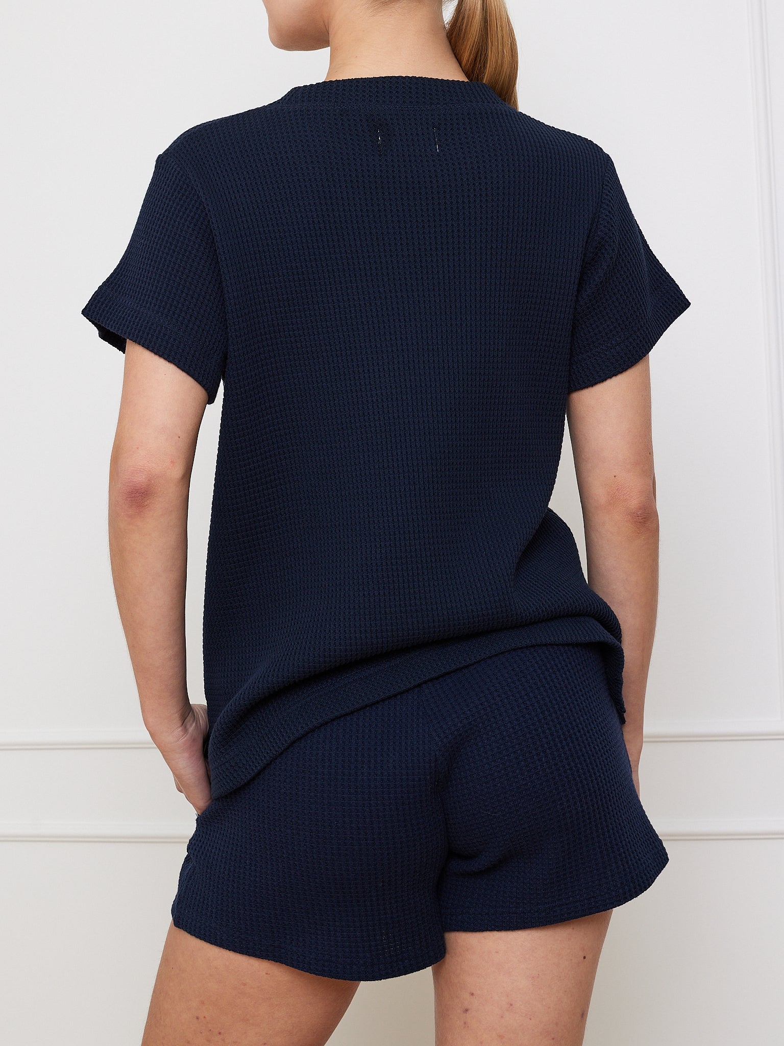 With their long hair tied back, a person wears a navy-blue Waffle Tee from We Norwegians and matching shorts. This wardrobe essential ensemble accentuates their silhouette as they stand with hands on hips against a white wall, facing away from the camera.