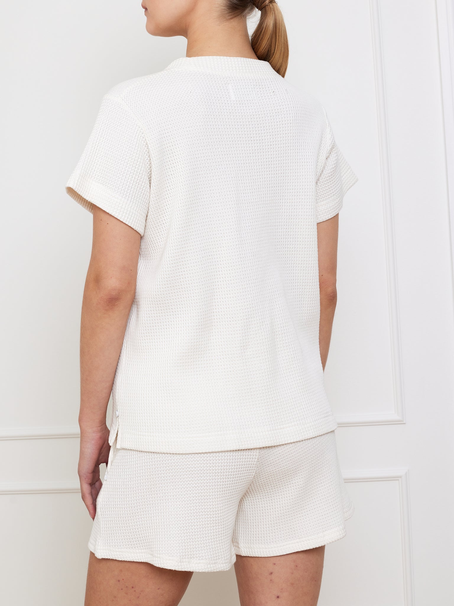 Rear view of a woman in a white Waffle Tee Women by We Norwegians and matching shorts, standing against a white paneled wall with her ponytail effortlessly pulled back. This waffle-knit piece is perfect for minimalist fashion lovers.