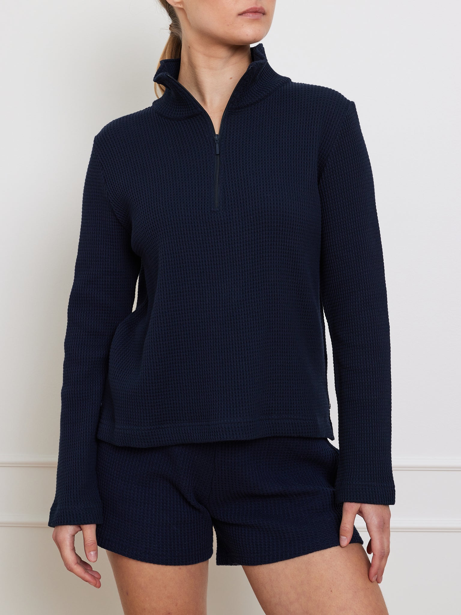 A person poses with hands at their sides, wearing the We Norwegians Waffle Zipped Sweater for women—a navy blue, relaxed fit, long-sleeve half-zip pullover with matching shorts—featuring a textured waffle knit pattern against a plain white background.