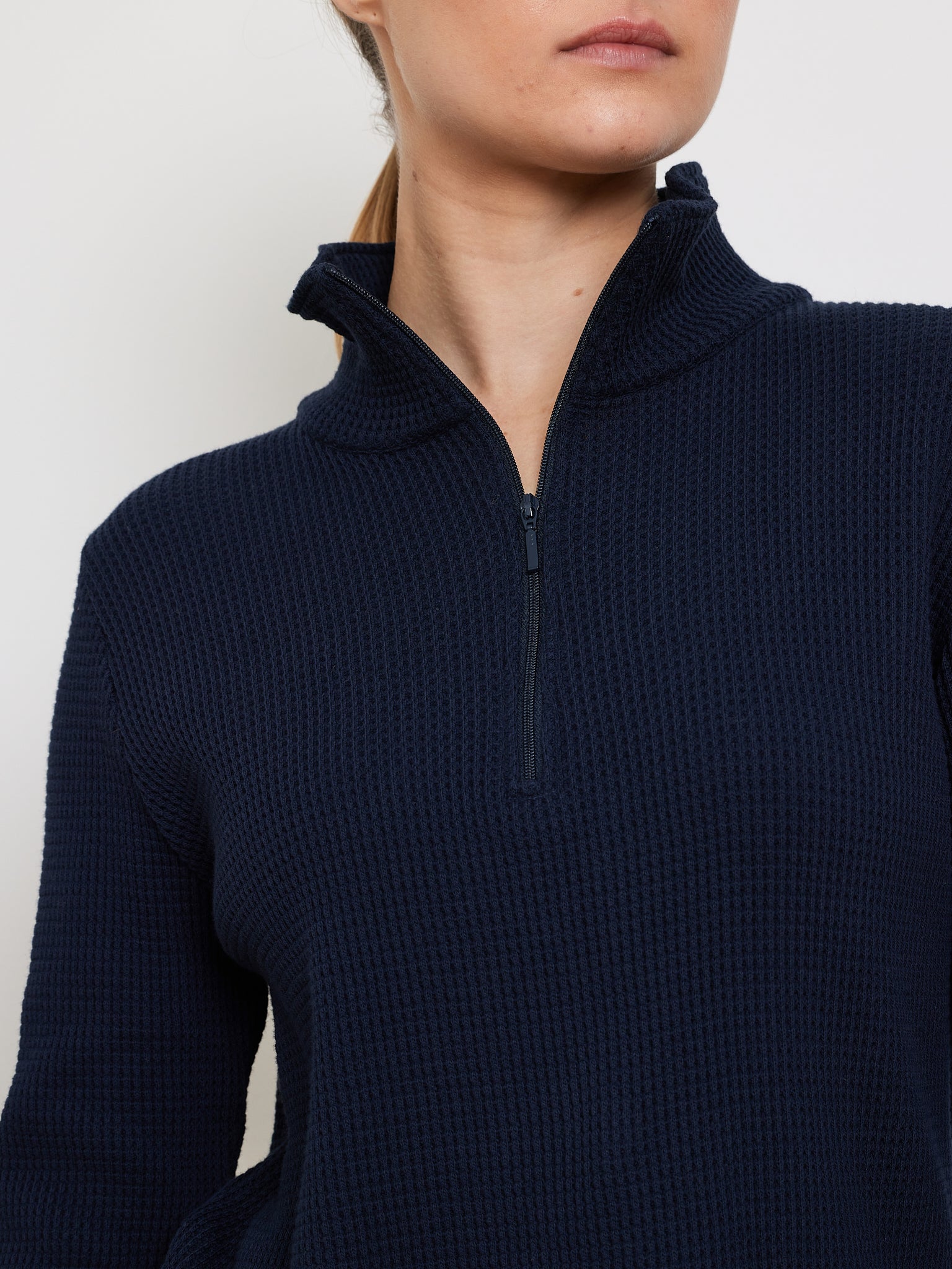A person models from shoulders down in a relaxed fit navy blue We Norwegians Waffle Zipped Sweater for women. The textured waffle knit adds depth, and the collar is partially zipped against a plain white backdrop.