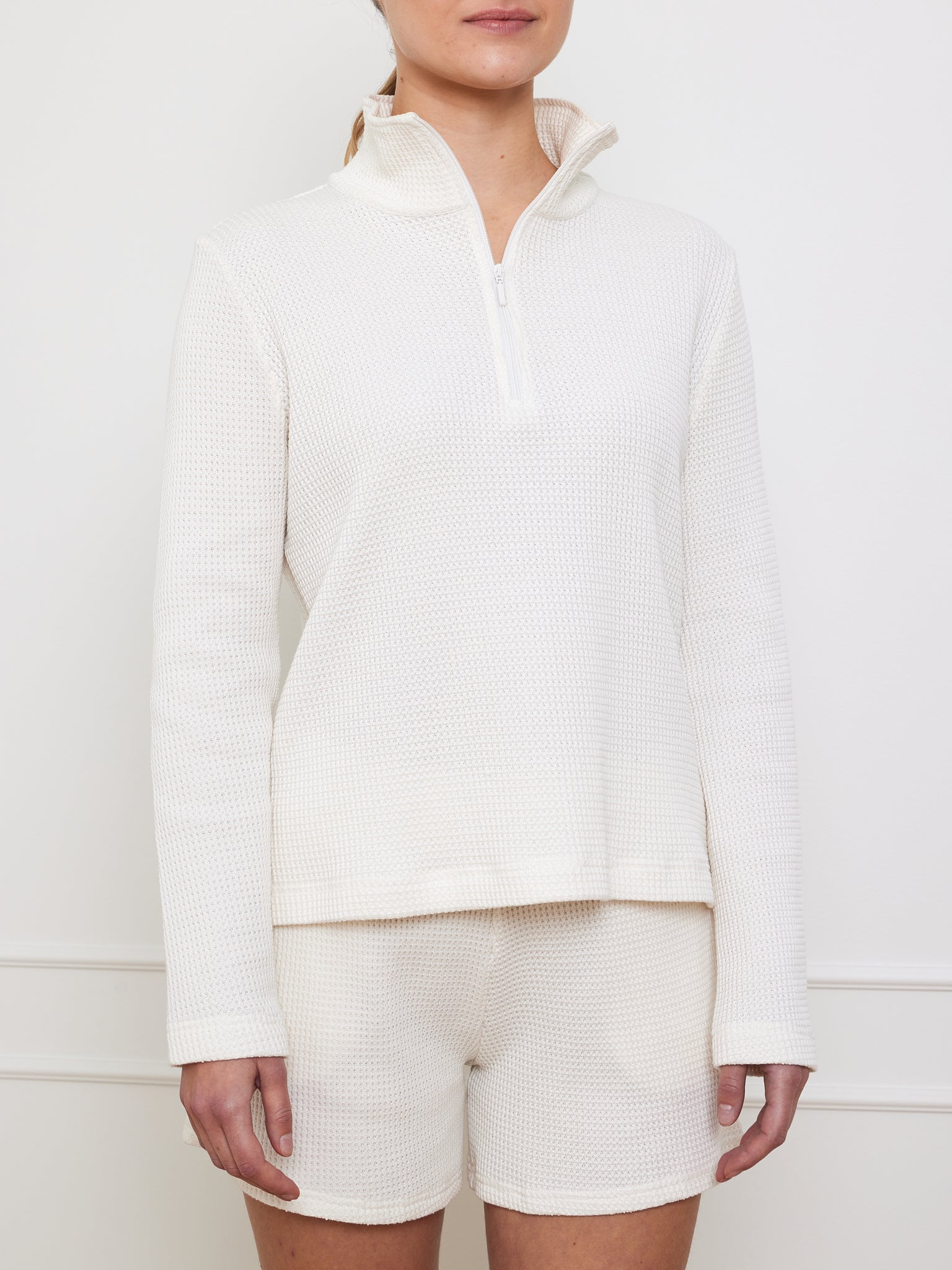 Dressed in We Norwegians' cream Waffle Zipped Sweater Women and matching shorts, a person showcases the textured waffle knit against a plain white wall; their head is not in the frame.