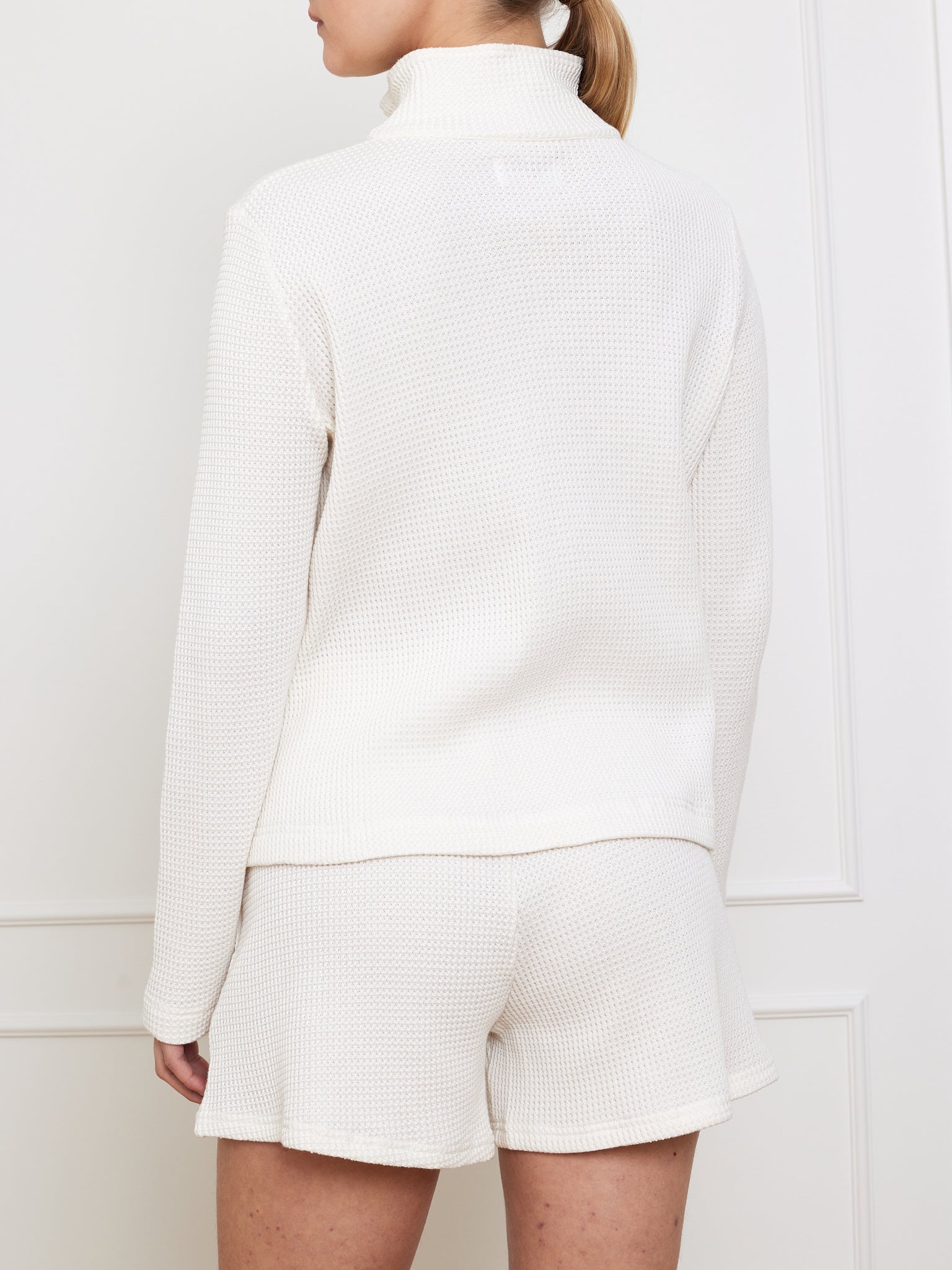 A person stands sideways against a white wall, wearing a relaxed-fit white waffle-textured long-sleeve top and matching shorts. The top, similar to the We Norwegians' Waffle Zipped Sweater Women, features a high collar. Their hair is tied back.