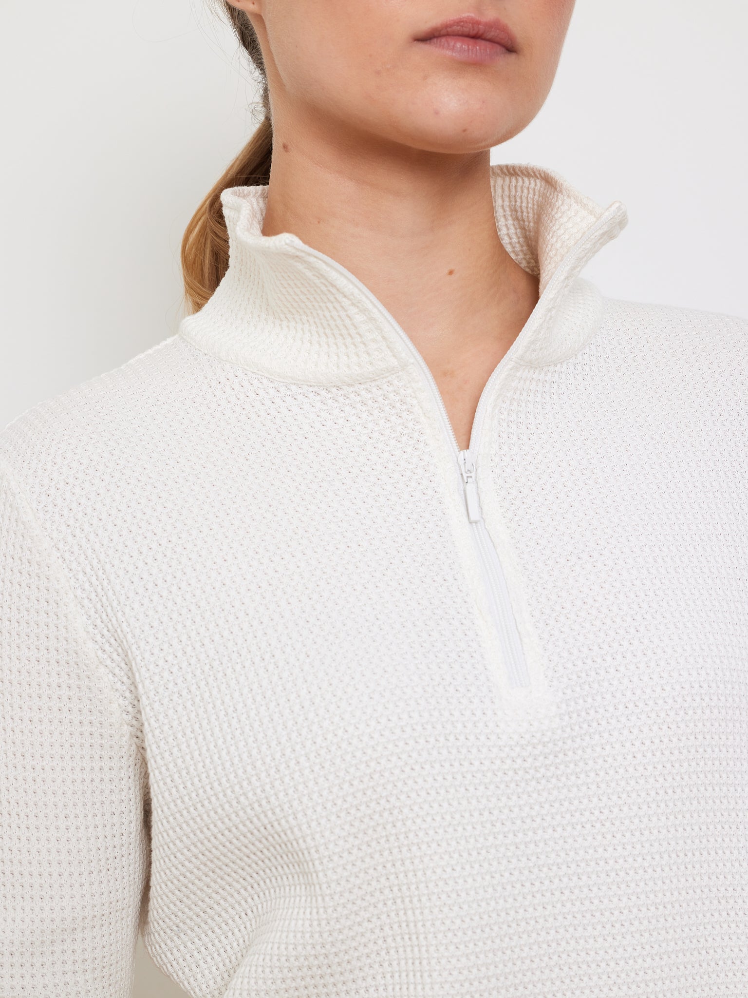 Close-up of someone in a relaxed-fit We Norwegians Waffle Zipped Sweater Women in white, showcasing a textured waffle knit and high collar with half-zip. Their hair is tied back, accentuating both the sweater's upper part and their lower face.
