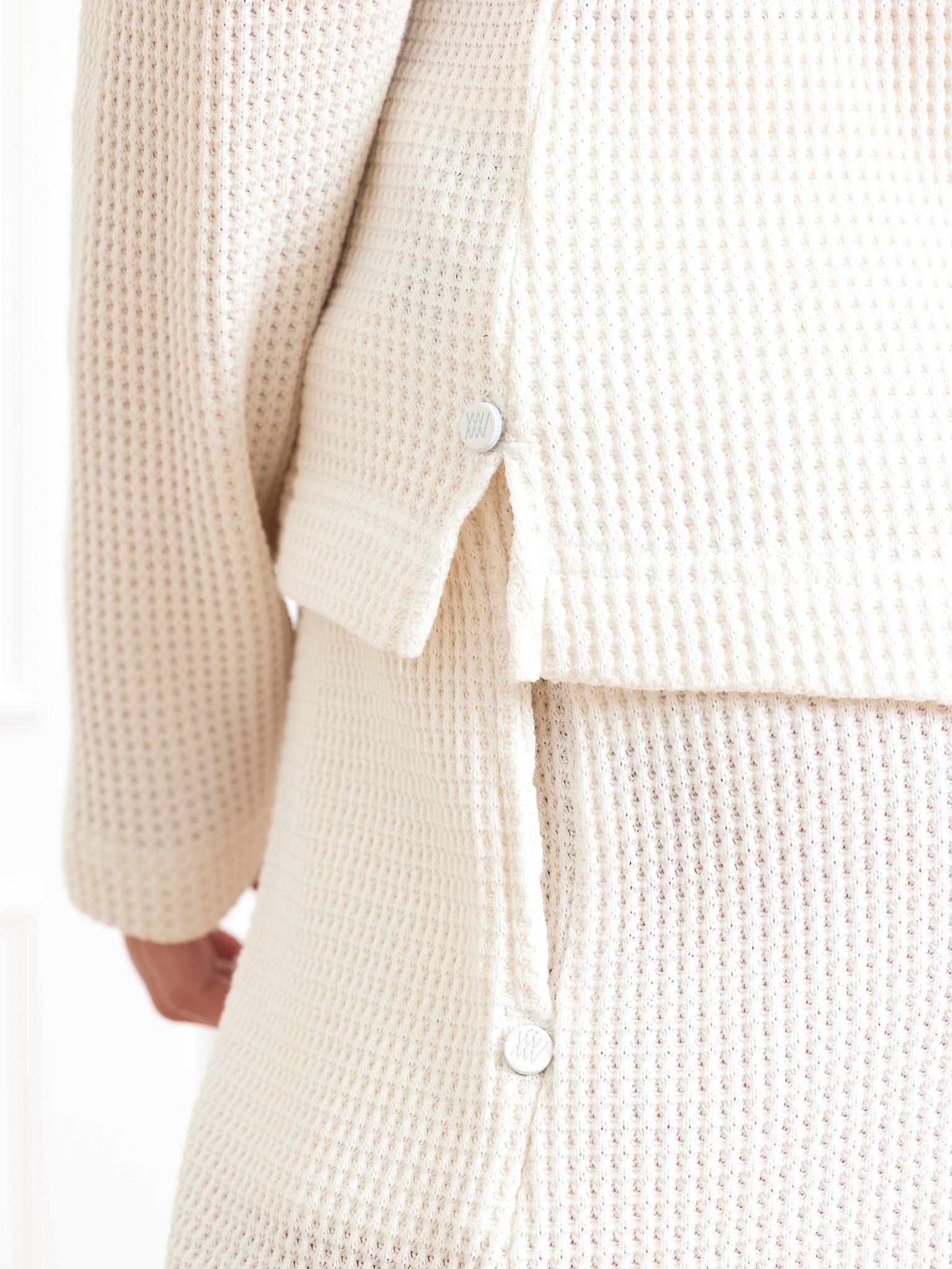 A person is wearing We Norwegians' Waffle Zipped Sweater Women, a cream-colored, waffle-knit loungewear set with a relaxed fit. The cozy and textured fabric makes it perfect for lounging, with side buttons on both the top and pants for easy adjustments.