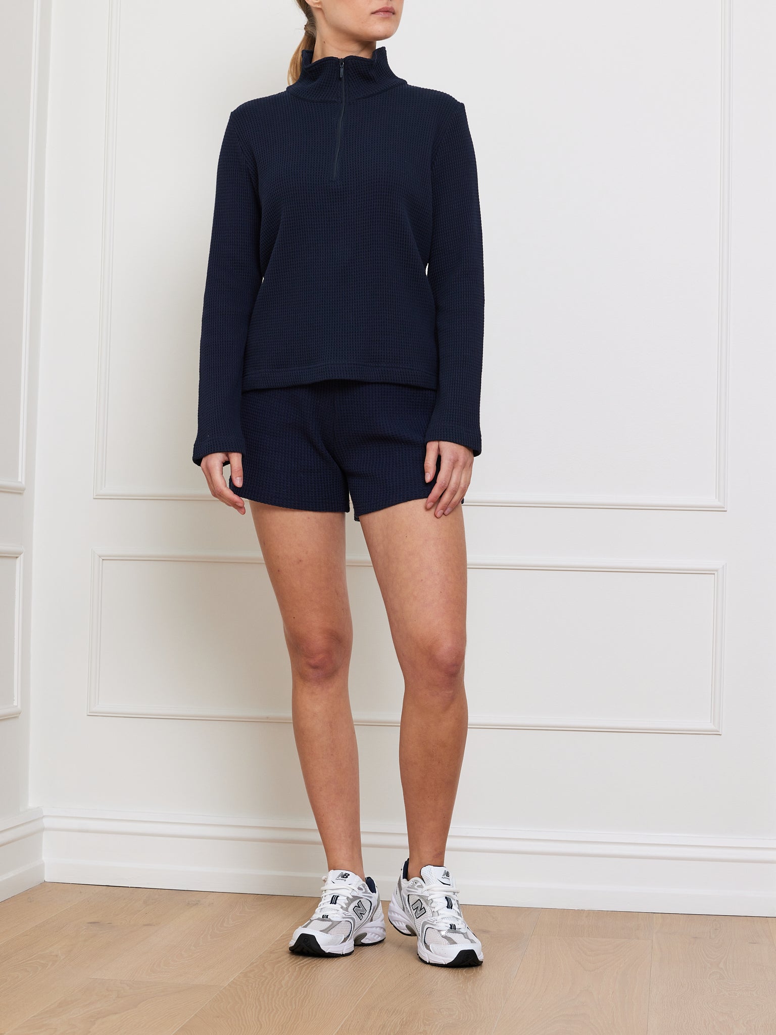 A person stands in a room wearing a navy blue long-sleeve quarter-zip top with We Norwegians' Waffle Shorts Women and white sneakers. The background's white paneled wall and light wood floor enhance the relaxed fit vibe of their outfit.