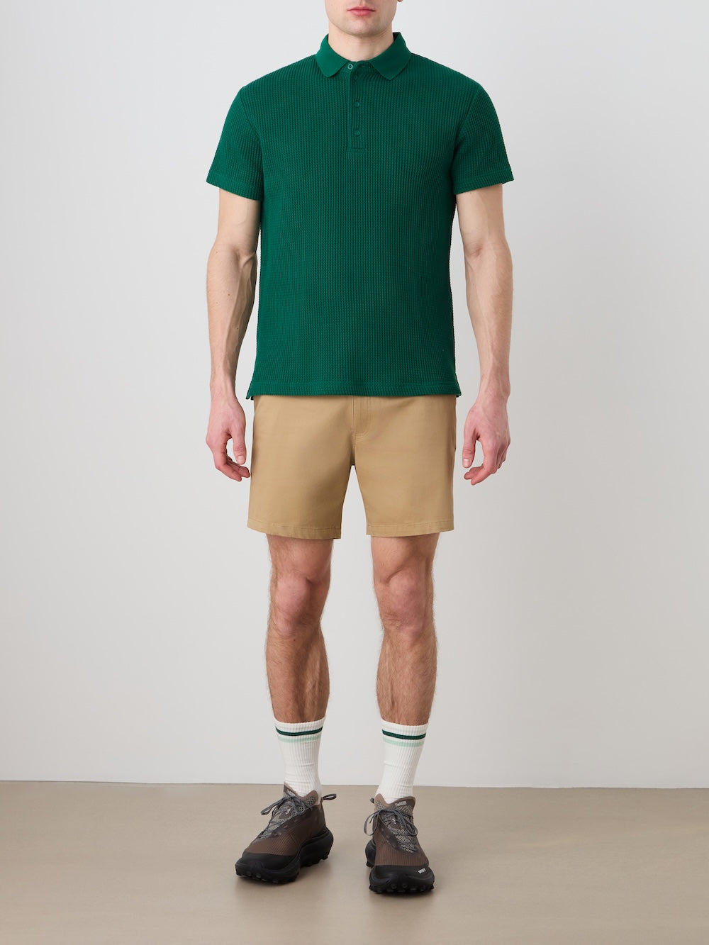A person stands against a plain background wearing the We Norwegians Waffle Polo Men, tan shorts, white socks with green stripes, and black shoes. The image is cropped at the neck, so the face is not visible.
