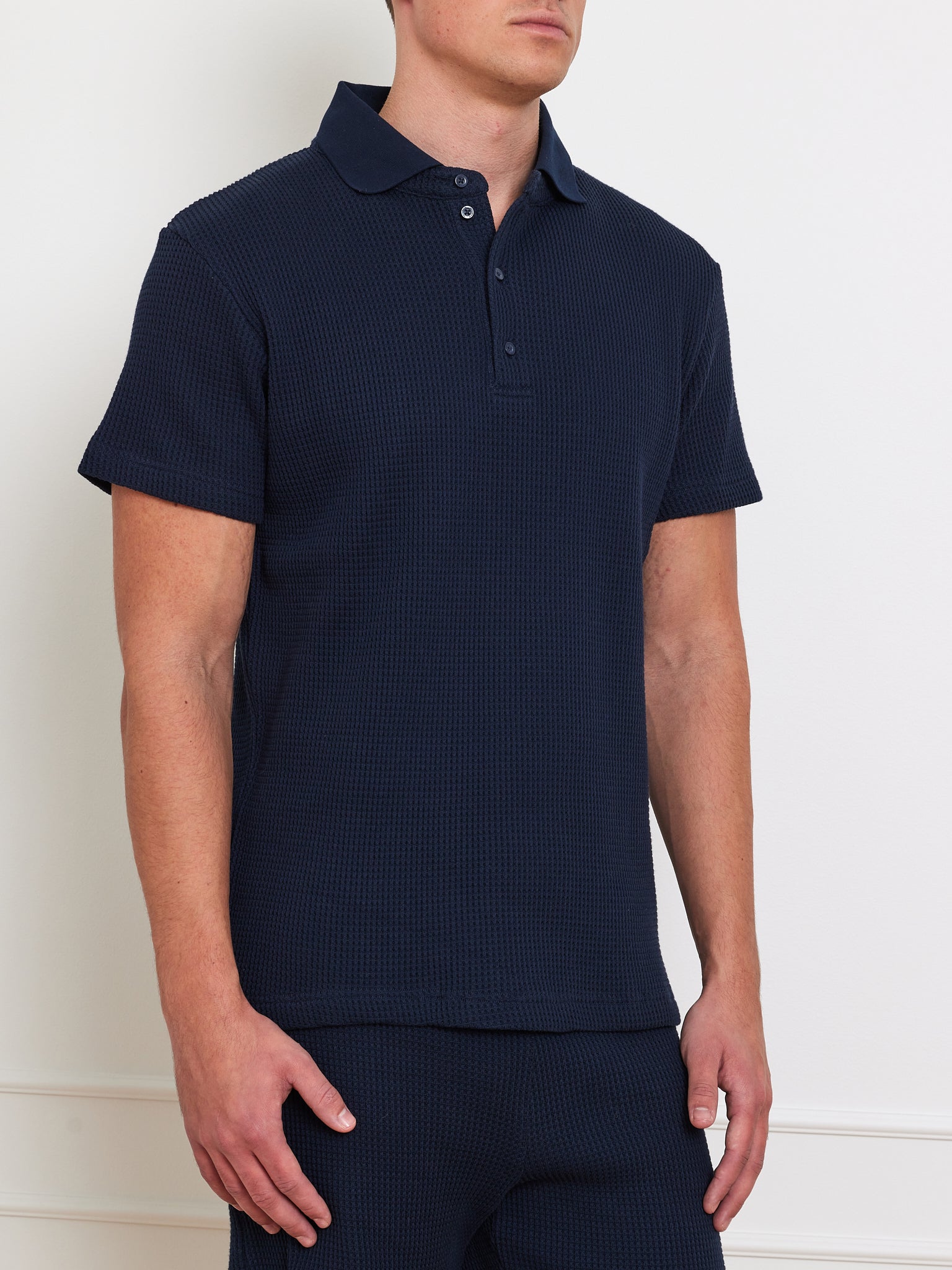 A man wearing We Norwegians' Waffle Polo Men, a textured waffle knit dark blue short-sleeve polo with button-up collar and relaxed fit, stands against a neutral background with his left hand casually by his side.