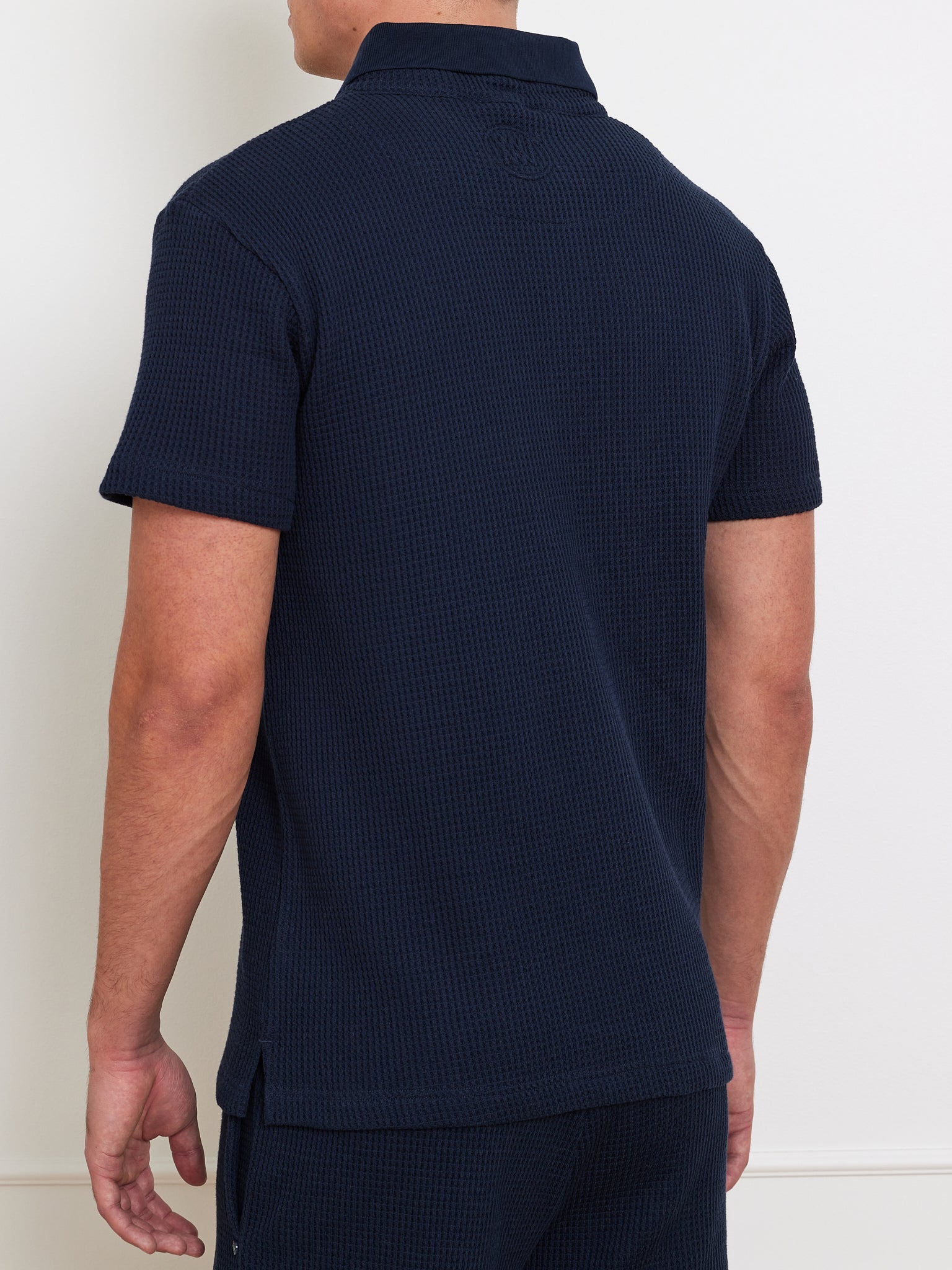 A man wearing a We Norwegians Waffle Polo Men in navy blue, noted for its relaxed fit, stands with his back turned. The shirt features a collared neck and short sleeves. He has short hair and is dressed in dark pants against a light-colored wall.
