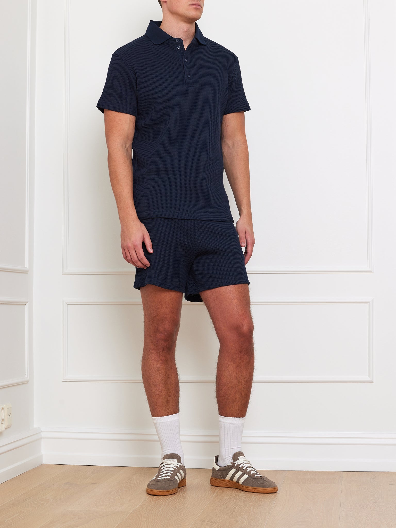 Dressed in a relaxed fit We Norwegians Waffle Polo Men and matching shorts, a person stands in a minimalist room. They wear brown athletic shoes with white stripes and long white socks. The white paneled walls and light wood flooring enhance the serene atmosphere.