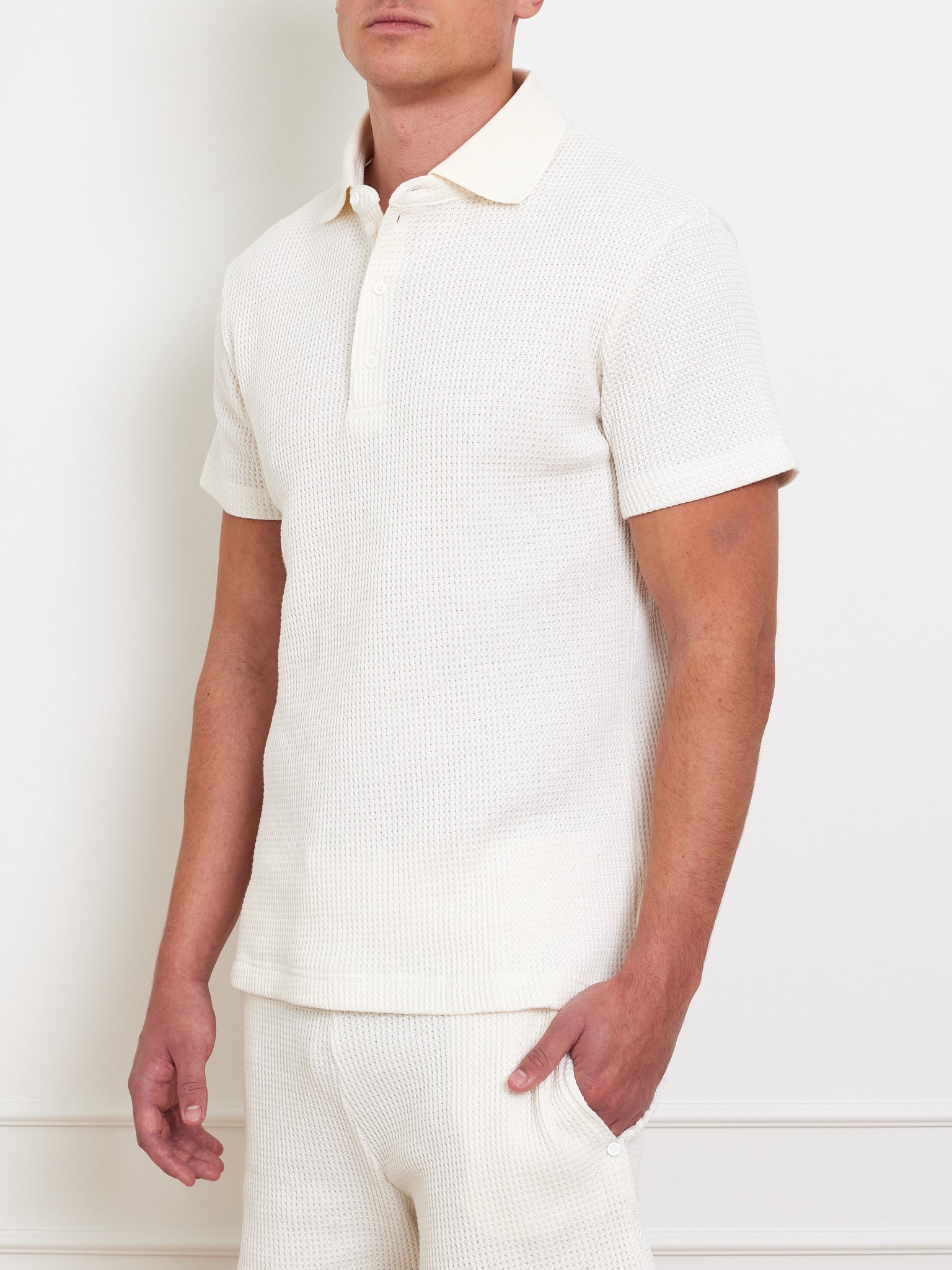 A person wears the "Waffle Polo Men" by We Norwegians, a white textured short-sleeve polo with a relaxed fit, and stands against a neutral background. Their head is slightly turned away from the camera, revealing a partial profile. They also wear matching white pants.