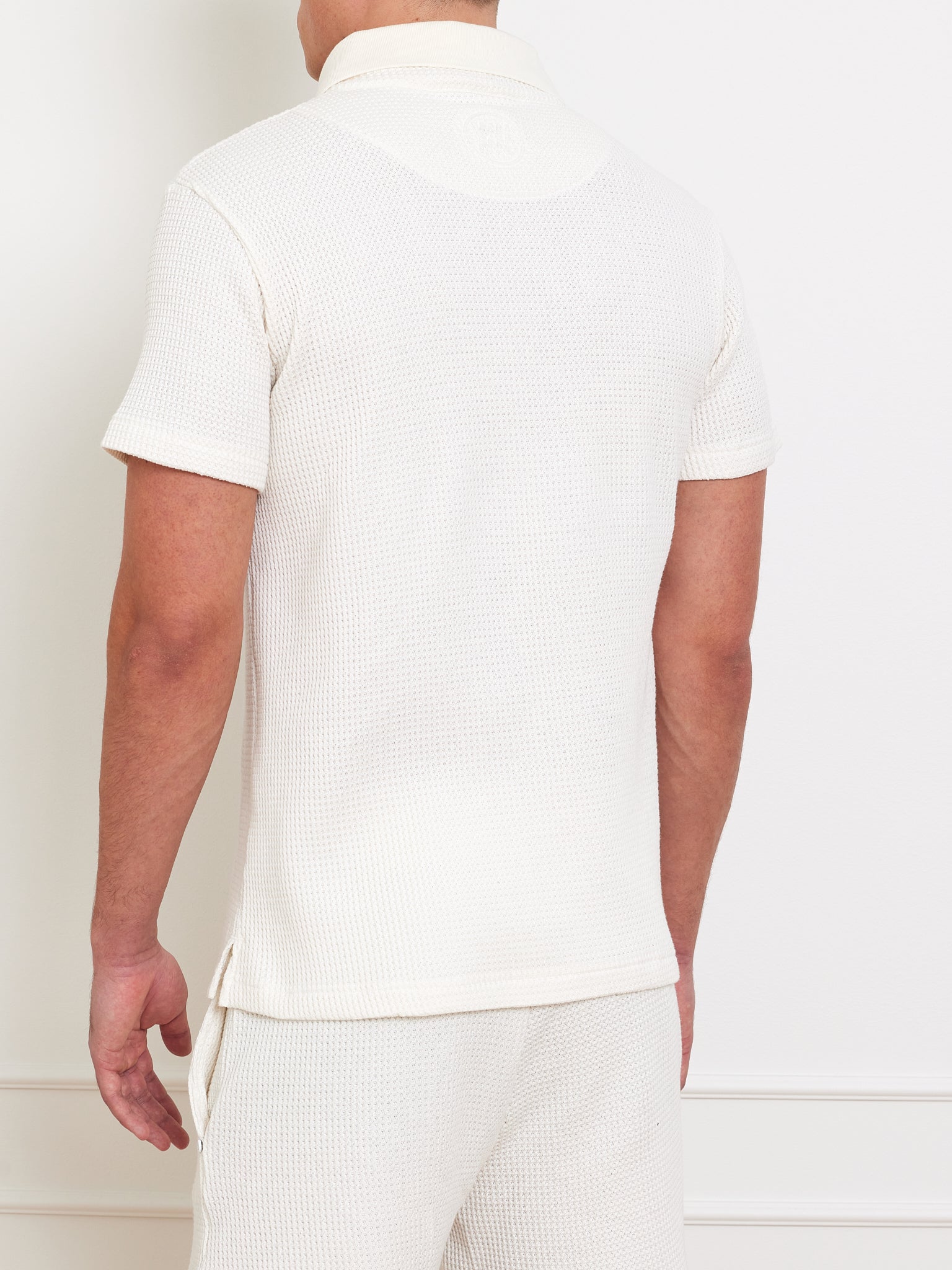A person in a We Norwegians Waffle Polo Men relaxed-fit, off-white short-sleeve shirt and matching pants stands with their back to the camera against a plain white background.