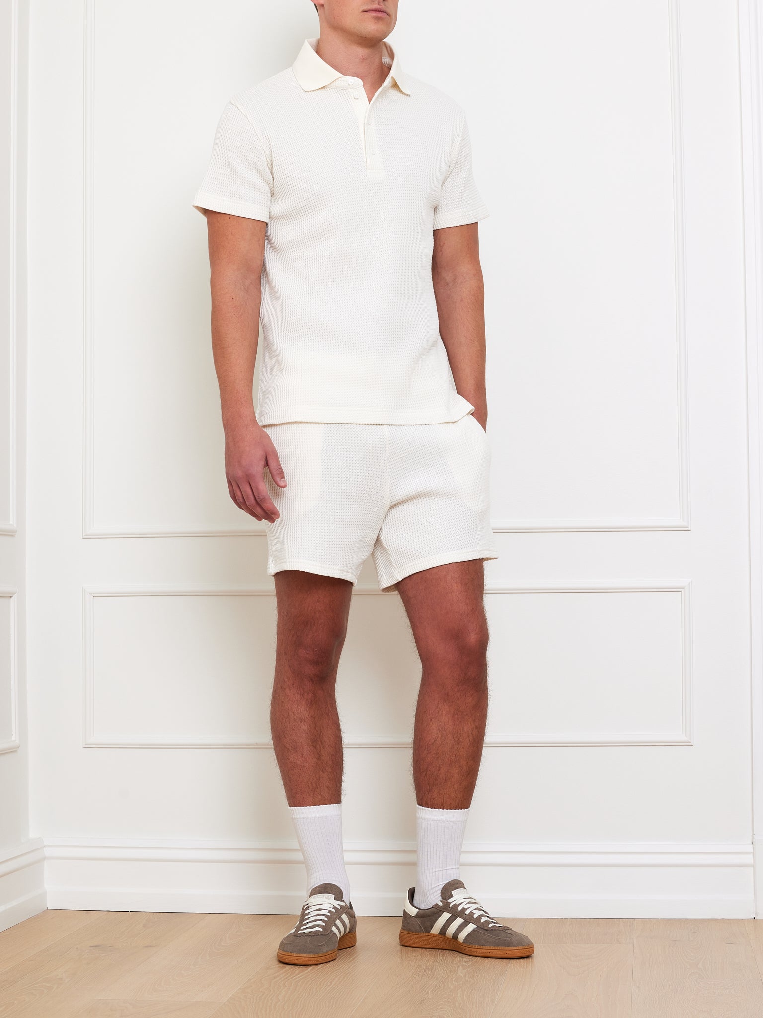 A person stands against a white paneled wall wearing We Norwegians' Waffle Polo Men in cream with matching relaxed-fit shorts. They complete the look with white socks and brown sneakers, but their face is cropped out.