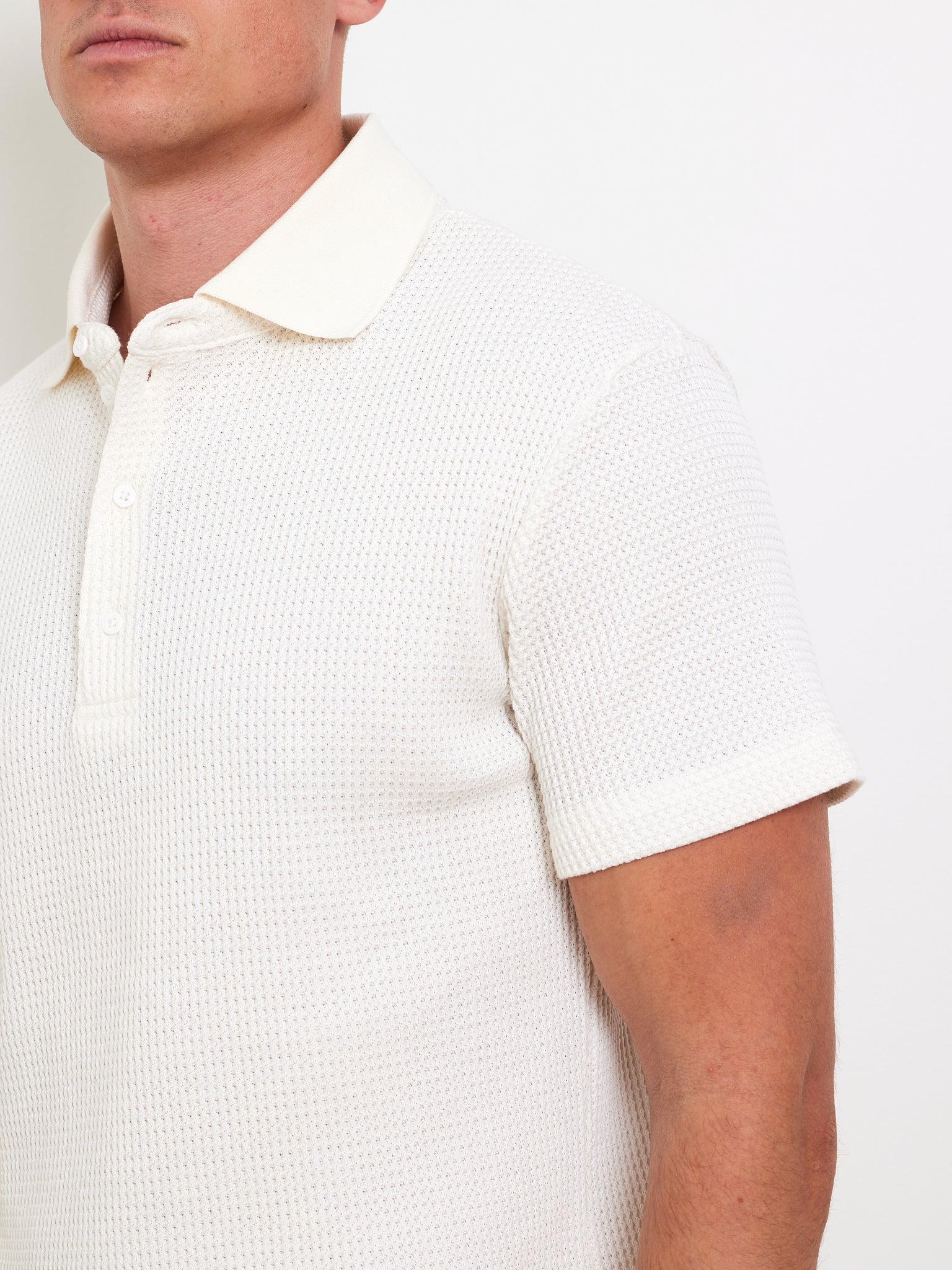 A man models the Waffle Polo Men by We Norwegians, featuring a textured white fabric, short sleeves, and a light-colored collar with buttons. This relaxed-fit piece stands out against a plain white backdrop, adding casual flair to the side-view upper body shot.