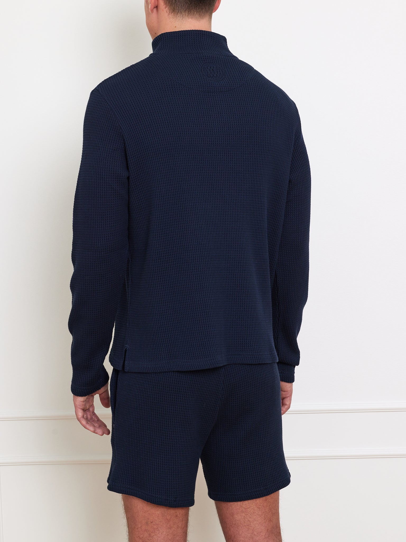 A person is wearing a Waffle Sweater Men by We Norwegians, made from dark navy, 100% cotton quilted fabric with long sleeves and a high neck. The outfit includes matching shorts, viewed from the back in a white room with minimal decor. They enjoy the relaxed fit.