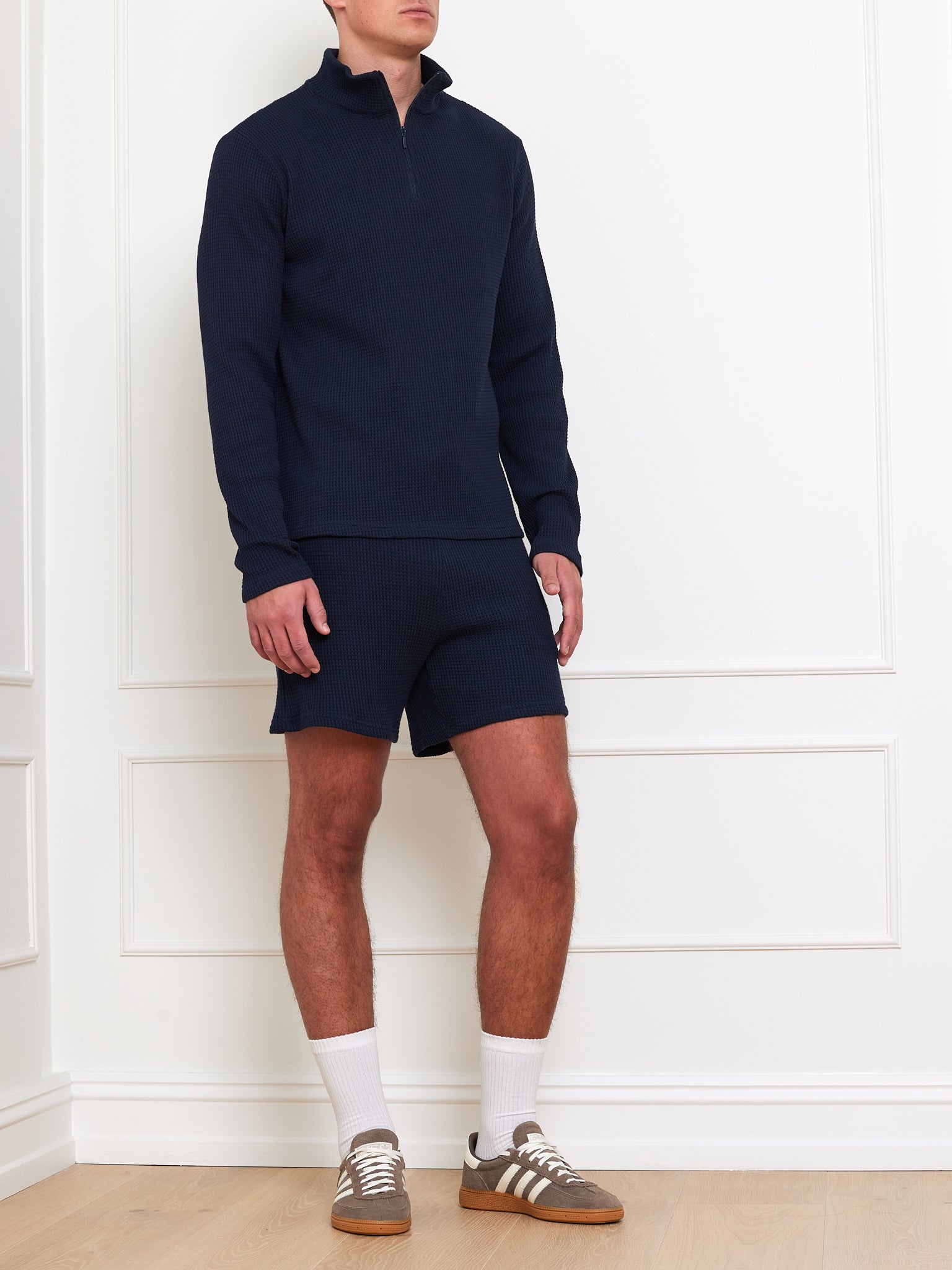 A man in a We Norwegians Waffle Sweater Men, zipped and in navy, teams it with matching shorts against a white wall. The look is completed with white socks and brown sneakers, effortlessly blending comfort and style.